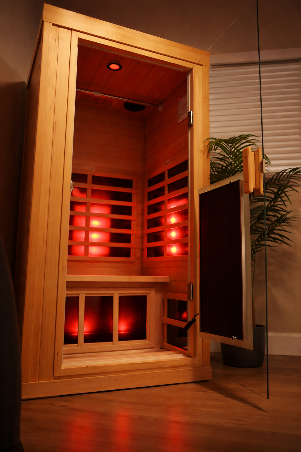 An infrared sauna with a wooden frame and door, featuring red lighting inside, located indoors next to a potted plant.