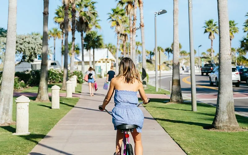 Woman riding a bicycle along a palm tree-lined sidewalk on a sunny day.