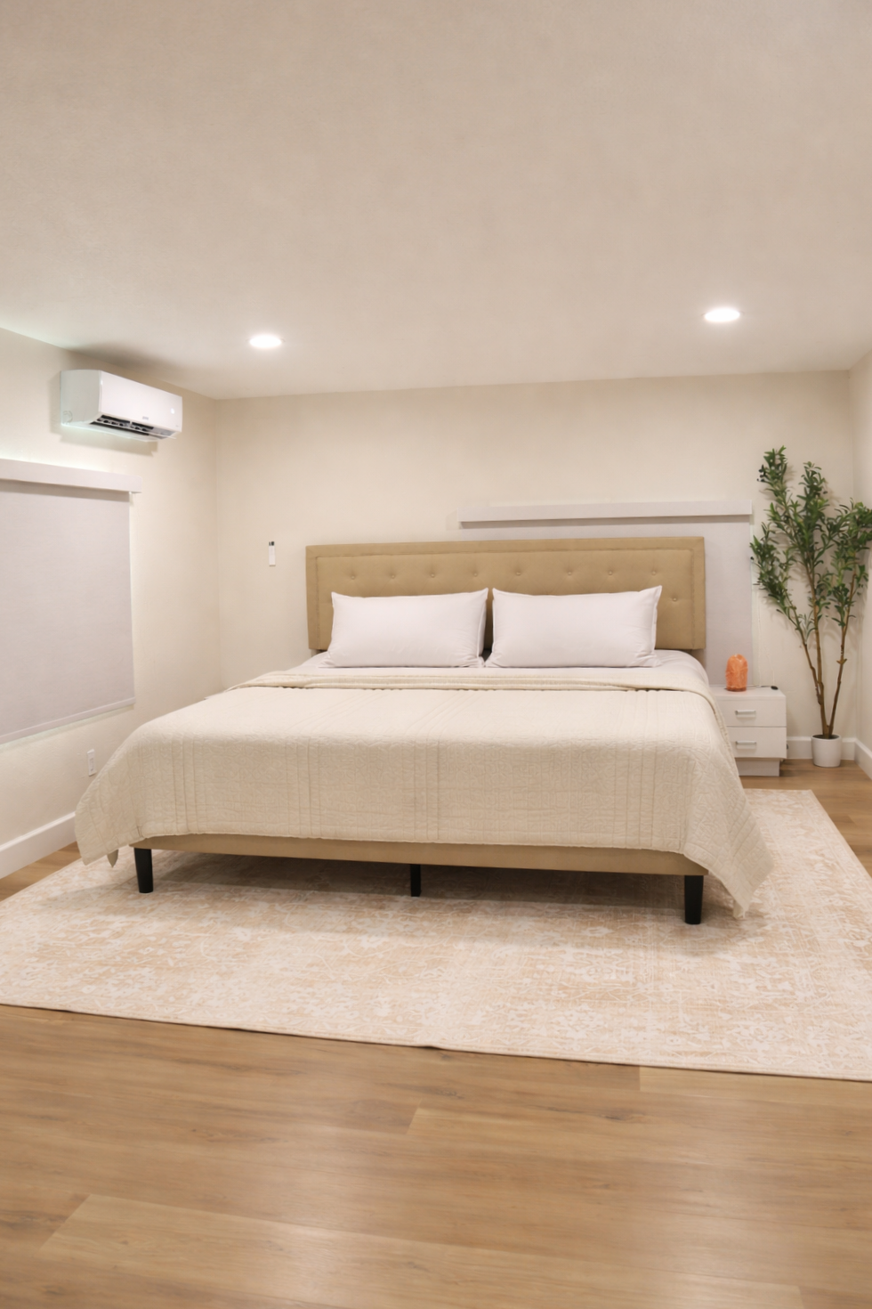 A bedroom with a king-sized bed featuring a beige upholstered headboard, white bedding, and three pillows. There is a white nightstand with a Himalayan salt lamp and a tall potted plant in the corner. The room has white walls, recessed lighting, a window with a blind, and a beige rug on wooden flooring.