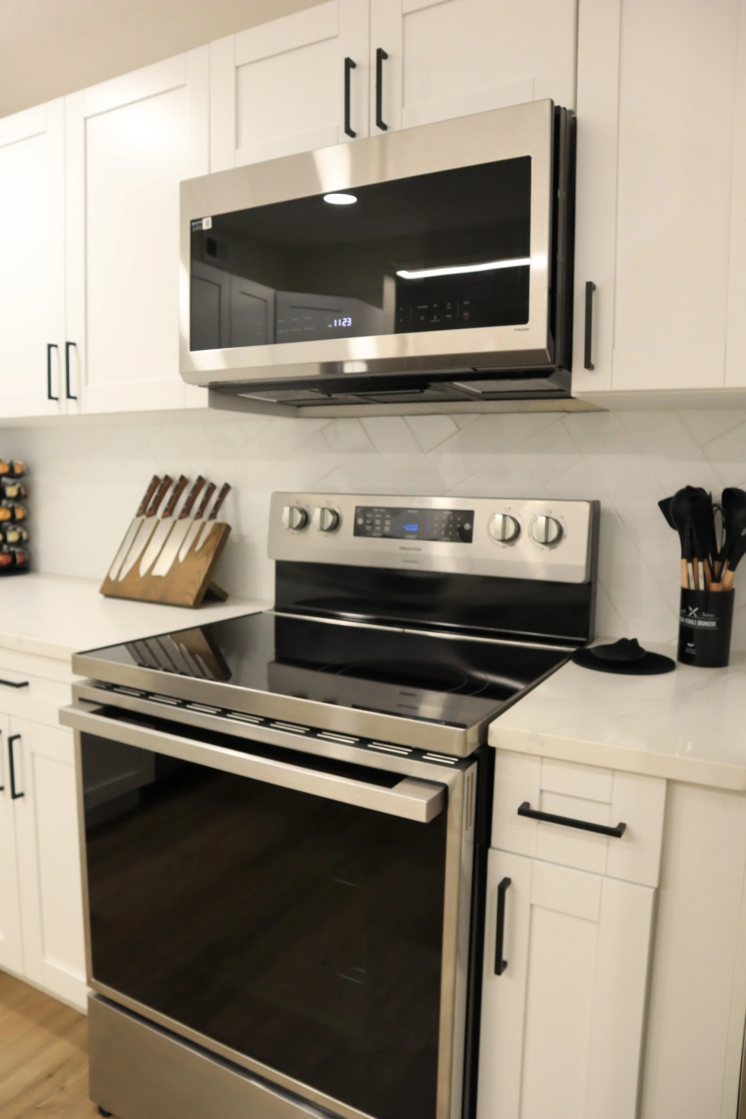 Stainless steel microwave oven installed above a stainless steel electric stove in a modern kitchen with white cabinetry, black handles, and a knife block on the countertop.