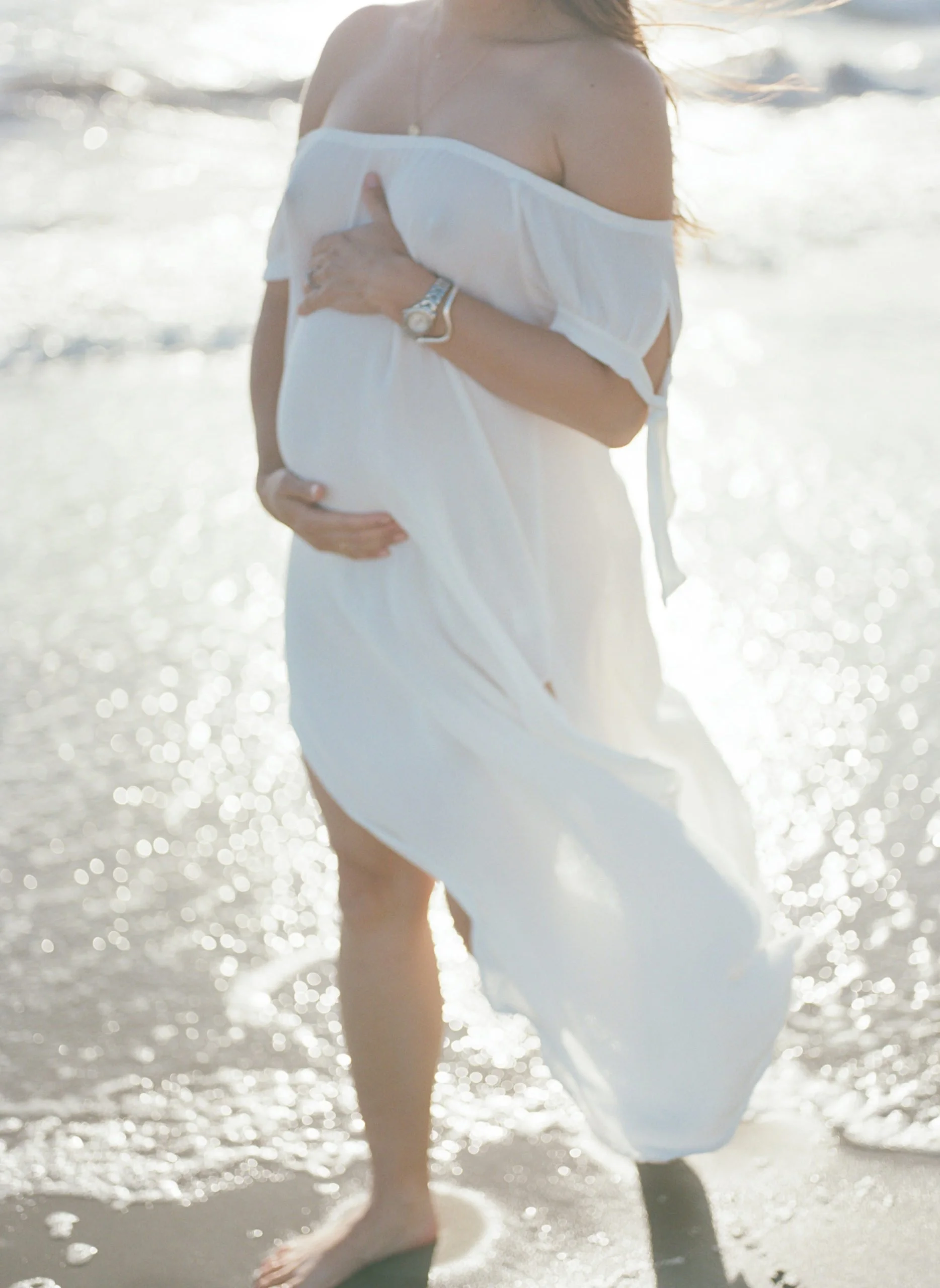 Pregnant woman in a white dress standing on a beach with water and sunlight in the background, holding her belly with both hands.