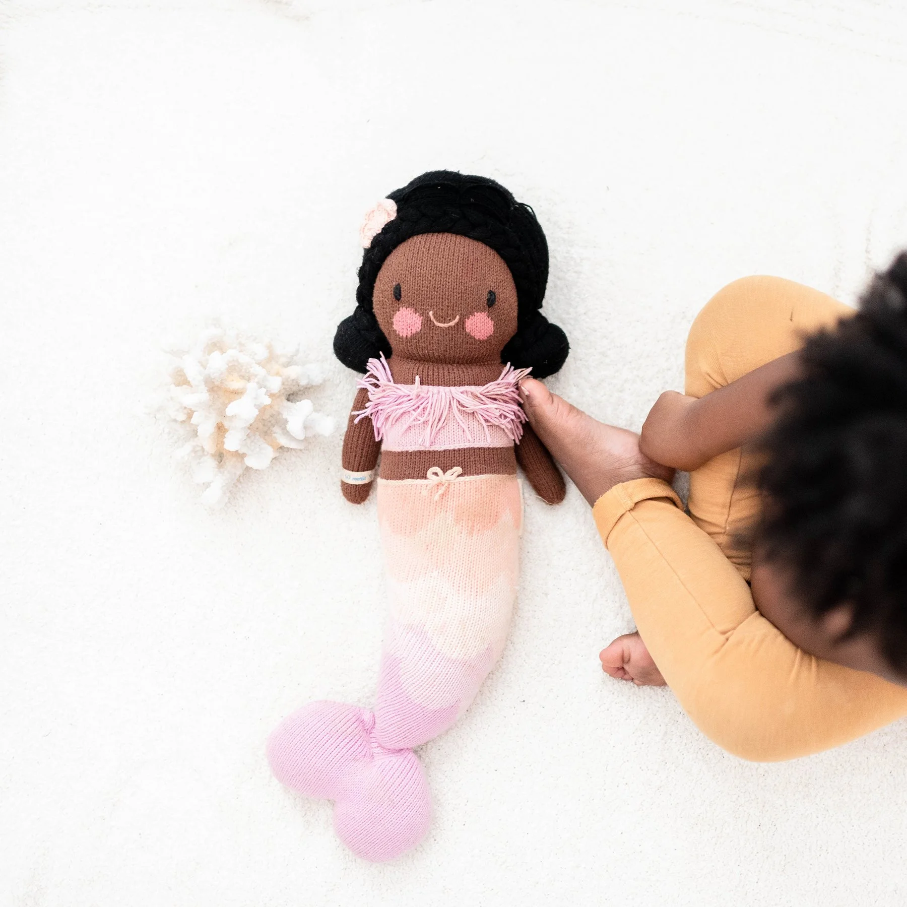 Child dressed in yellow pants holds a plush mermaid doll with brown skin, black hair, pink top, and pastel pink tail on a white surface.
