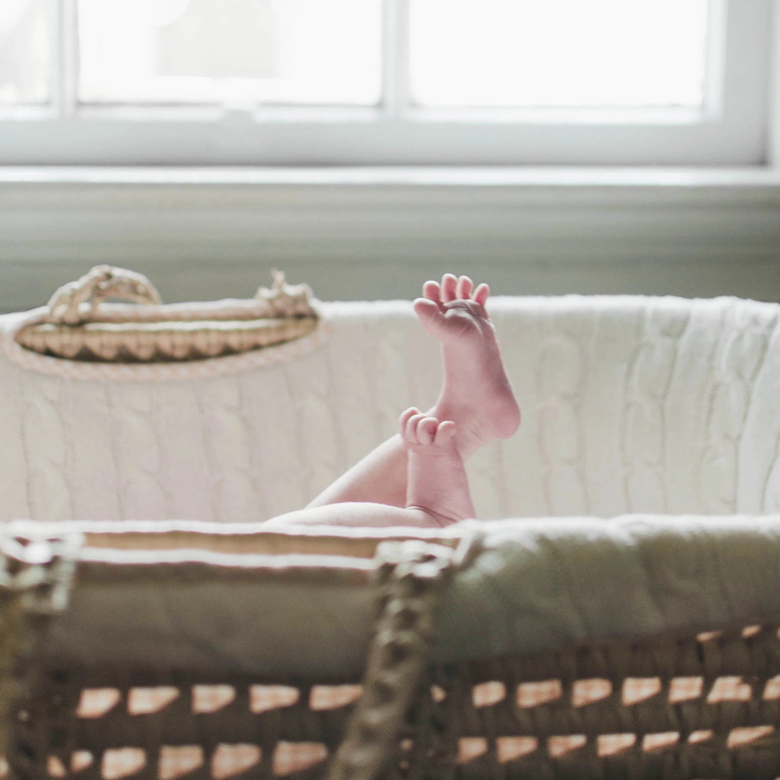 Close-up of a baby's legs and feet in a wicker bassinet near a window.