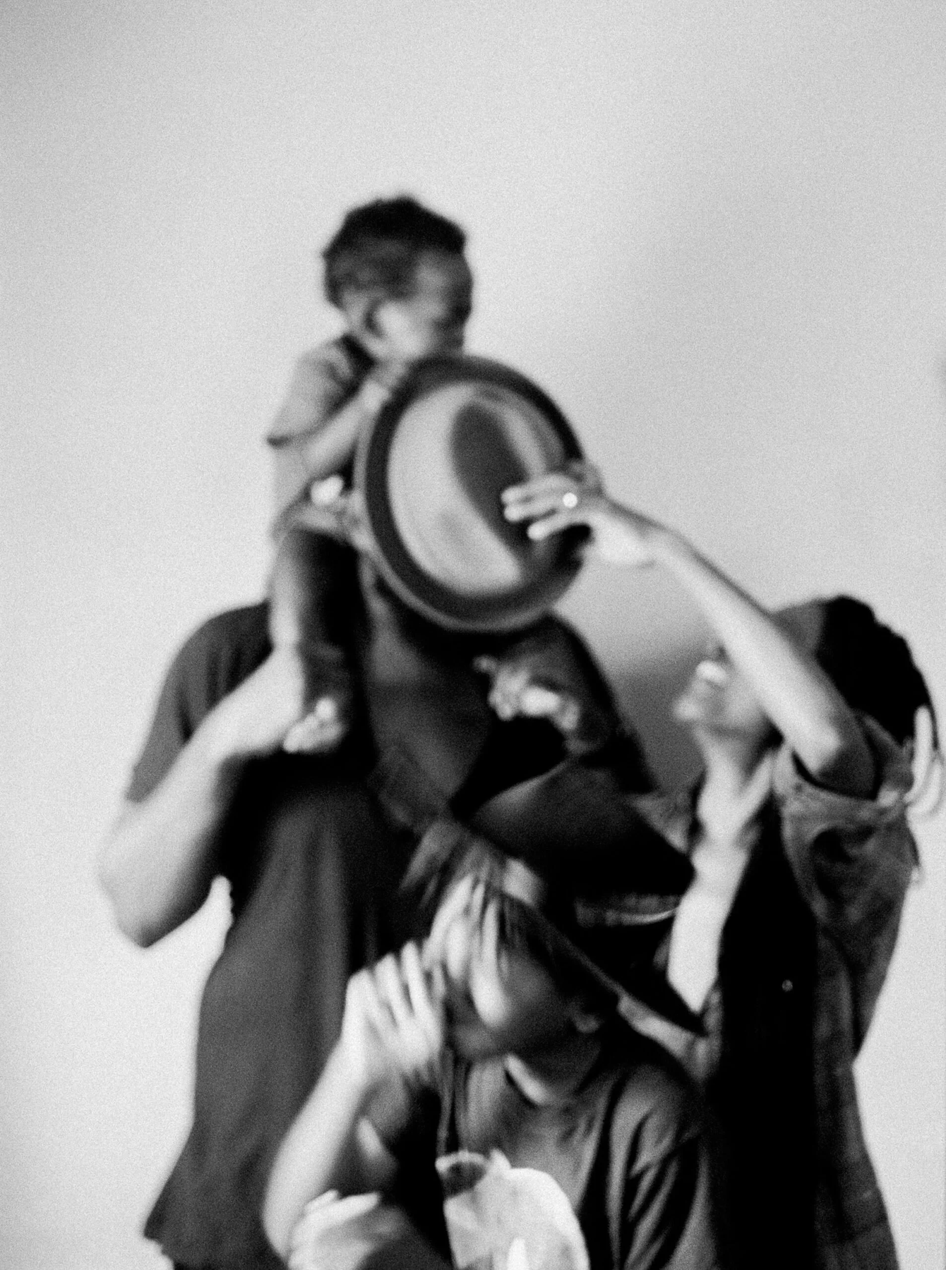 A person with glasses is carrying a child on their shoulders, and the child is holding a hat in front of the person's face. Another person is reaching up, touching the child's face, and smiling.