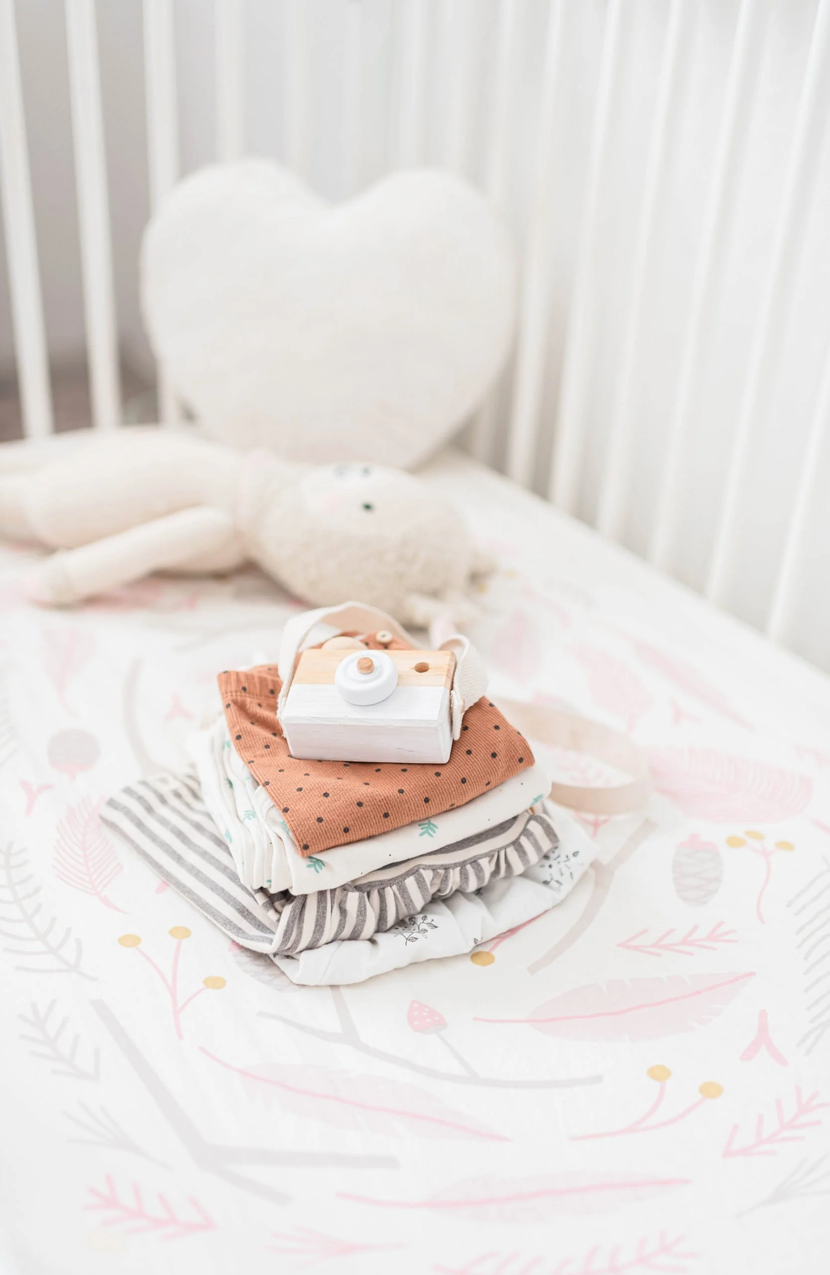 Stack of folded baby clothes on a crib sheet with stuffed animals and a heart-shaped pillow in the background.