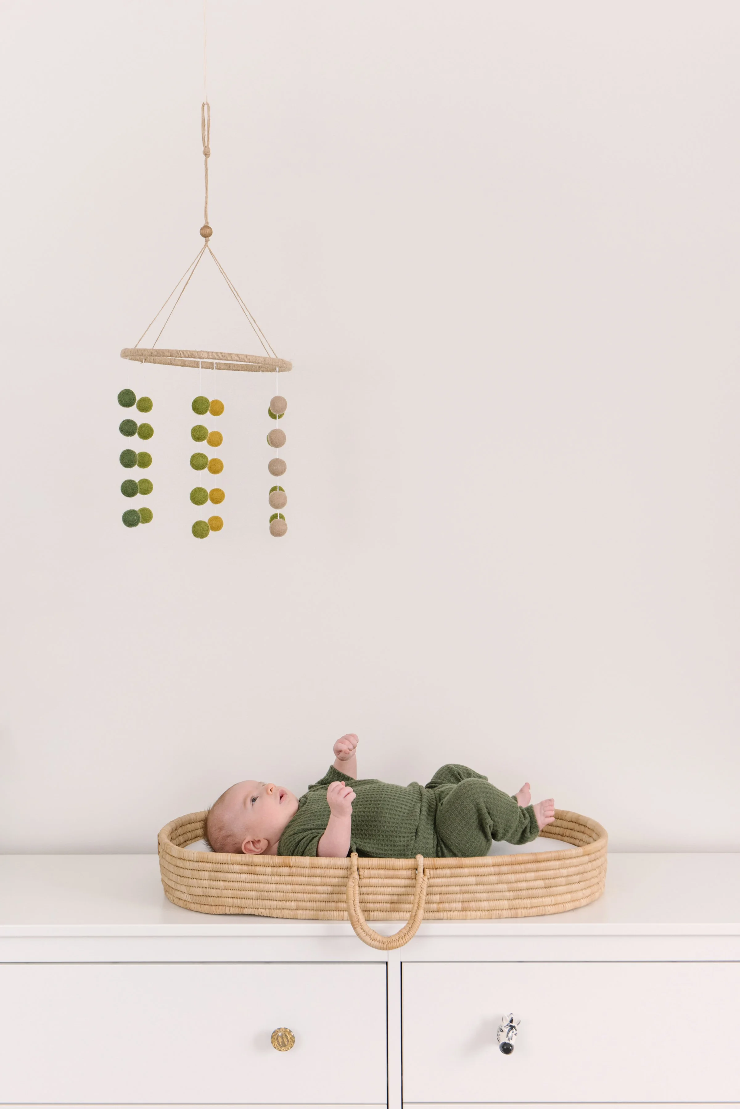 A baby lying in a woven basket on a white dresser, looking up at a hanging mobile with felt balls in green, yellow, and beige. The background is a plain white wall.