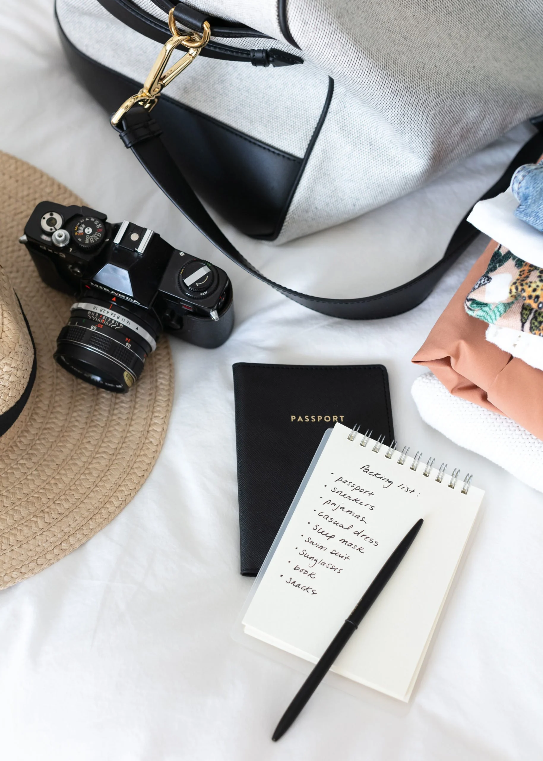 Travel essentials including a camera, a straw hat, a passport, a packing list, a notebook, and folded clothes on a white surface.