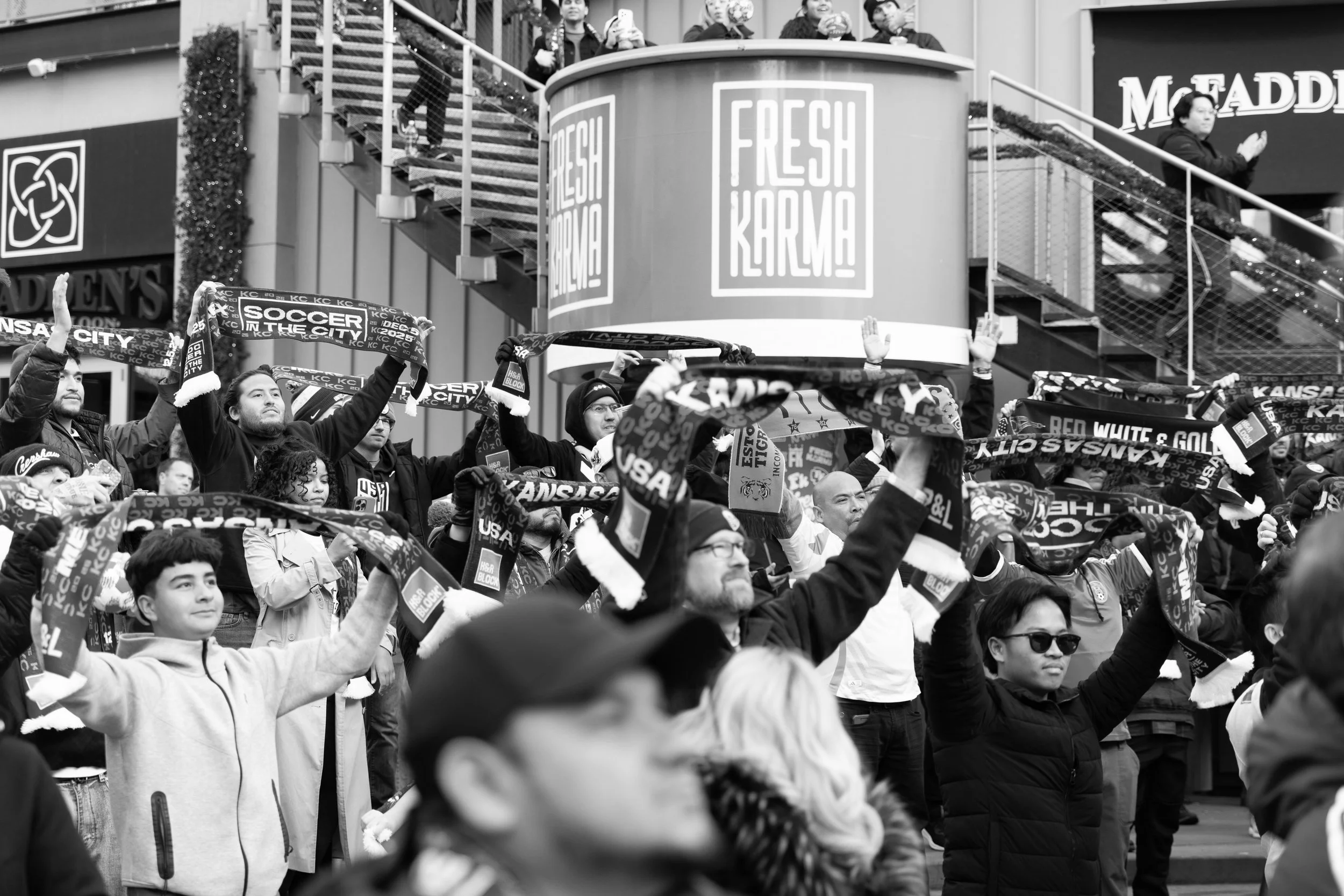 Crowd of people holding scarves and standing at a public event, some with raised hands, in front of stores and banners.