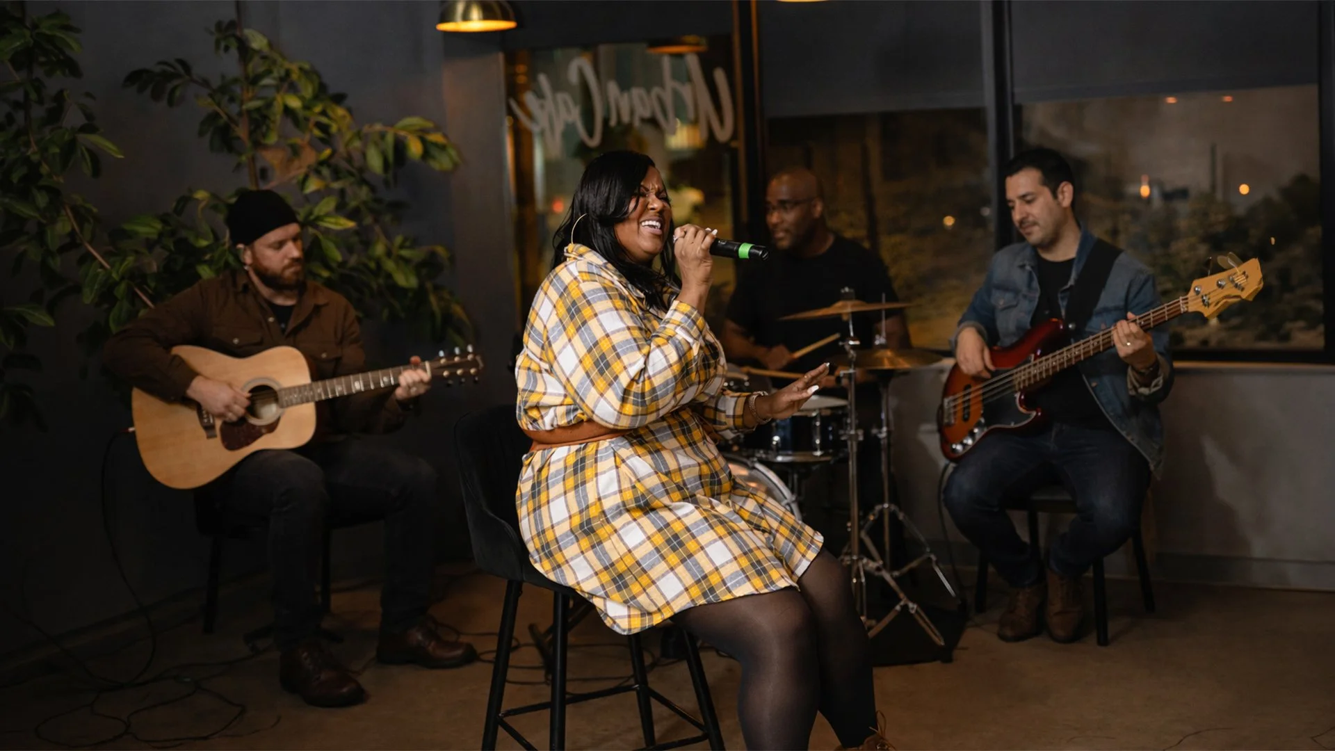 A woman in a yellow plaid dress singing into a microphone, accompanied by three men playing musical instruments in a cozy indoor setting with large windows and warm lighting.