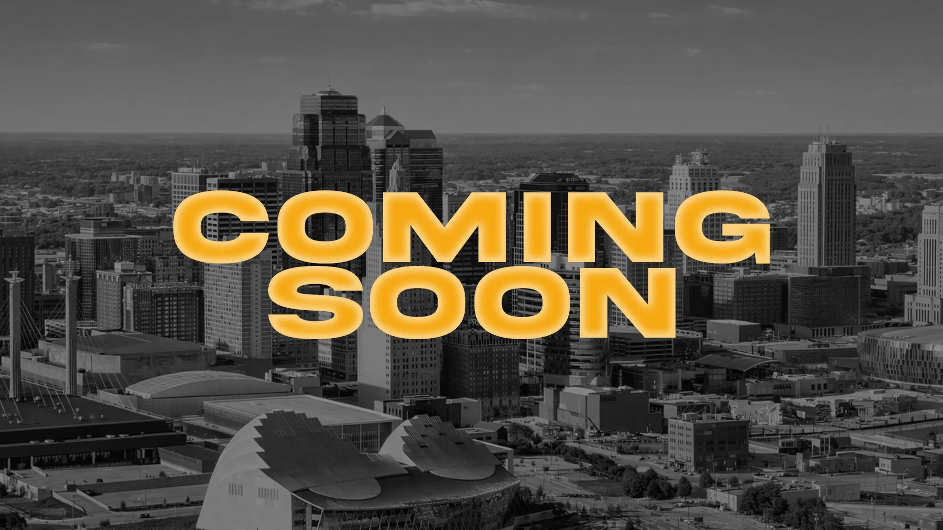 A black and white aerial view of a city skyline with tall buildings. Overlaid on the cityscape are large, bold, yellow-orange letters spelling out "COMING SOON."