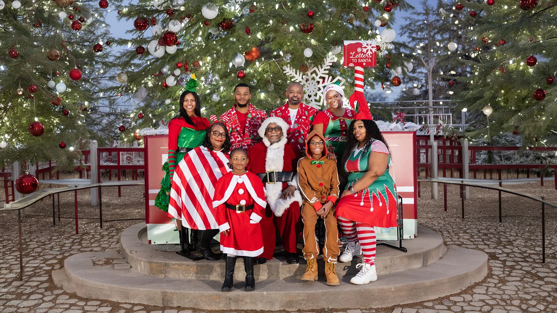 Group of people dressed in Christmas costumes and accessories posing in front of decorated Christmas trees with ornaments and lights, on outdoor steps with a sign that reads 'Letters to Santa'.