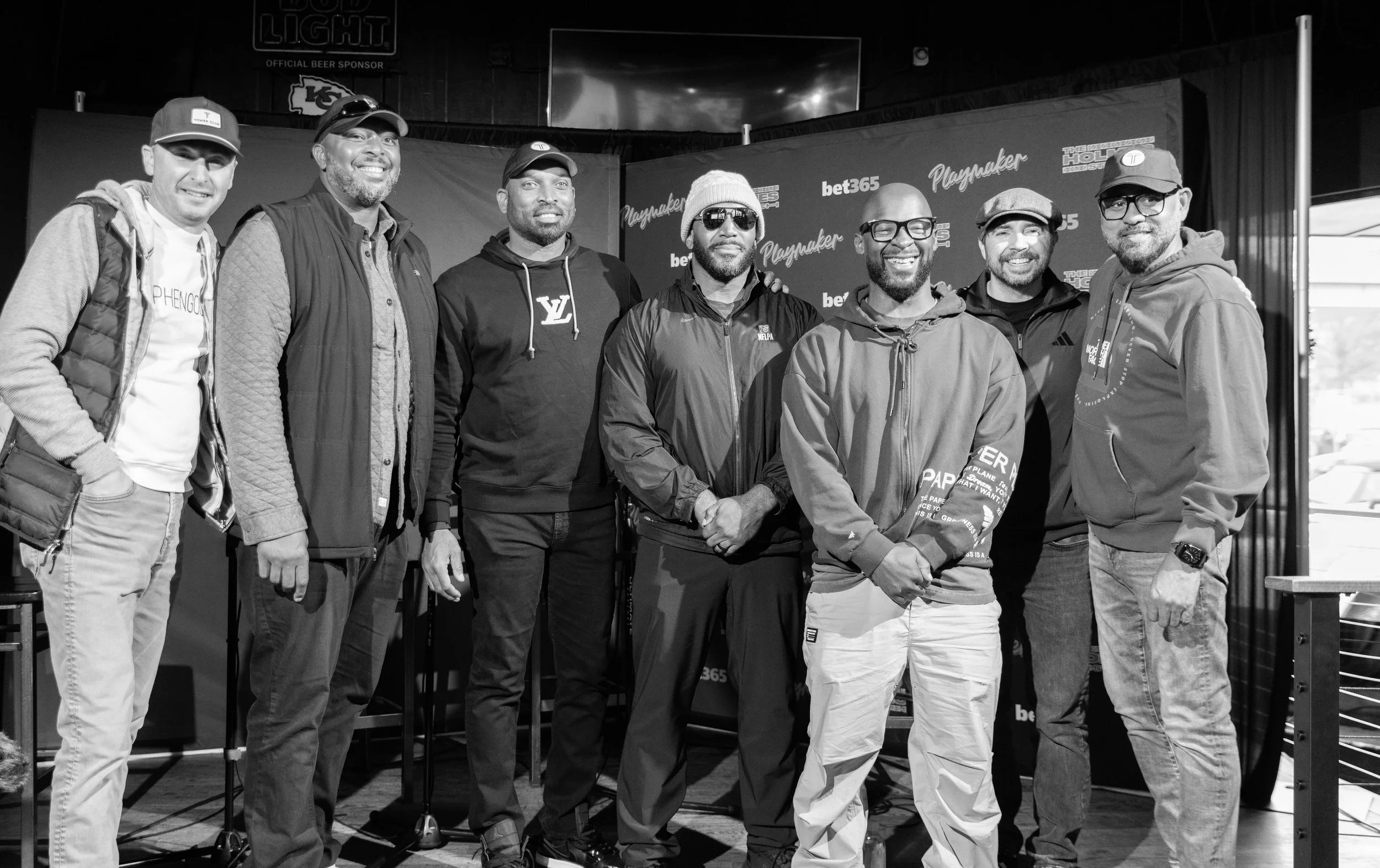 A group of eight men standing together on a stage or in a venue, smiling for the photo. They are dressed casually, some wearing jackets, hoodies, and caps. There is a backdrop behind them with logos and text, indicating a promotional or sponsored eve