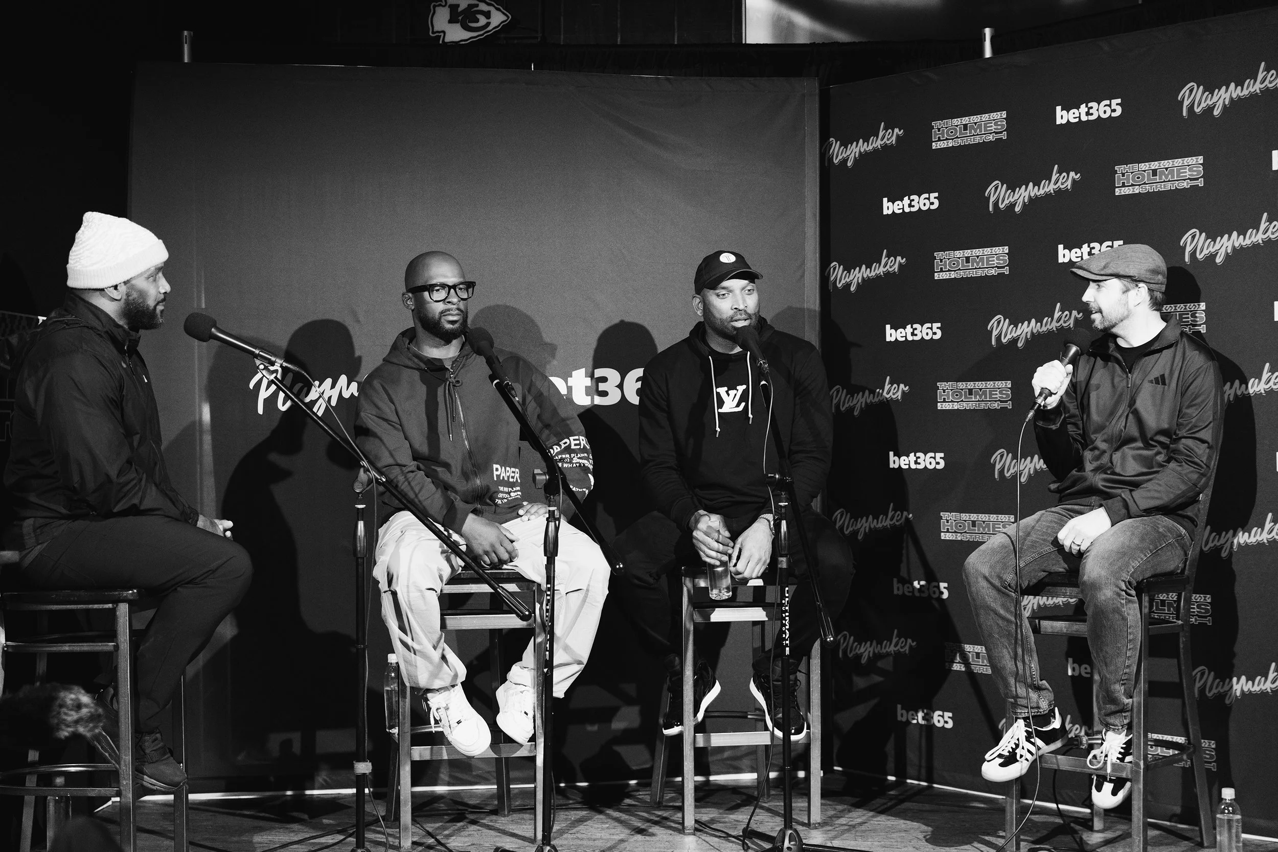 Four men sitting on stools and talking into microphones during a panel discussion at an event with a backdrop featuring logos of Playmaker, Bet365, Holmes, and Louis Vuitton.