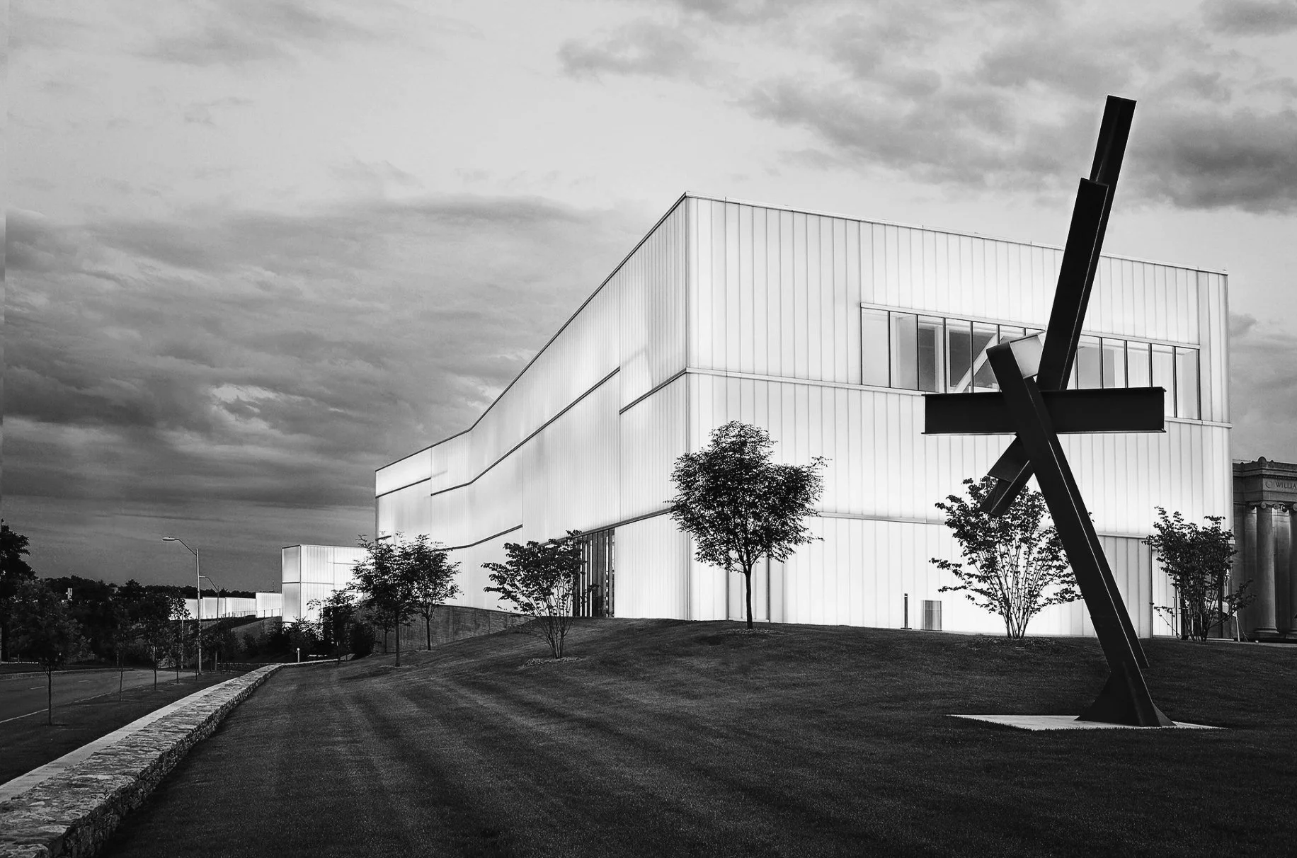 Modern white building with angular design, large windows, trees in front, and abstract black sculpture on lawn, under cloudy sky.