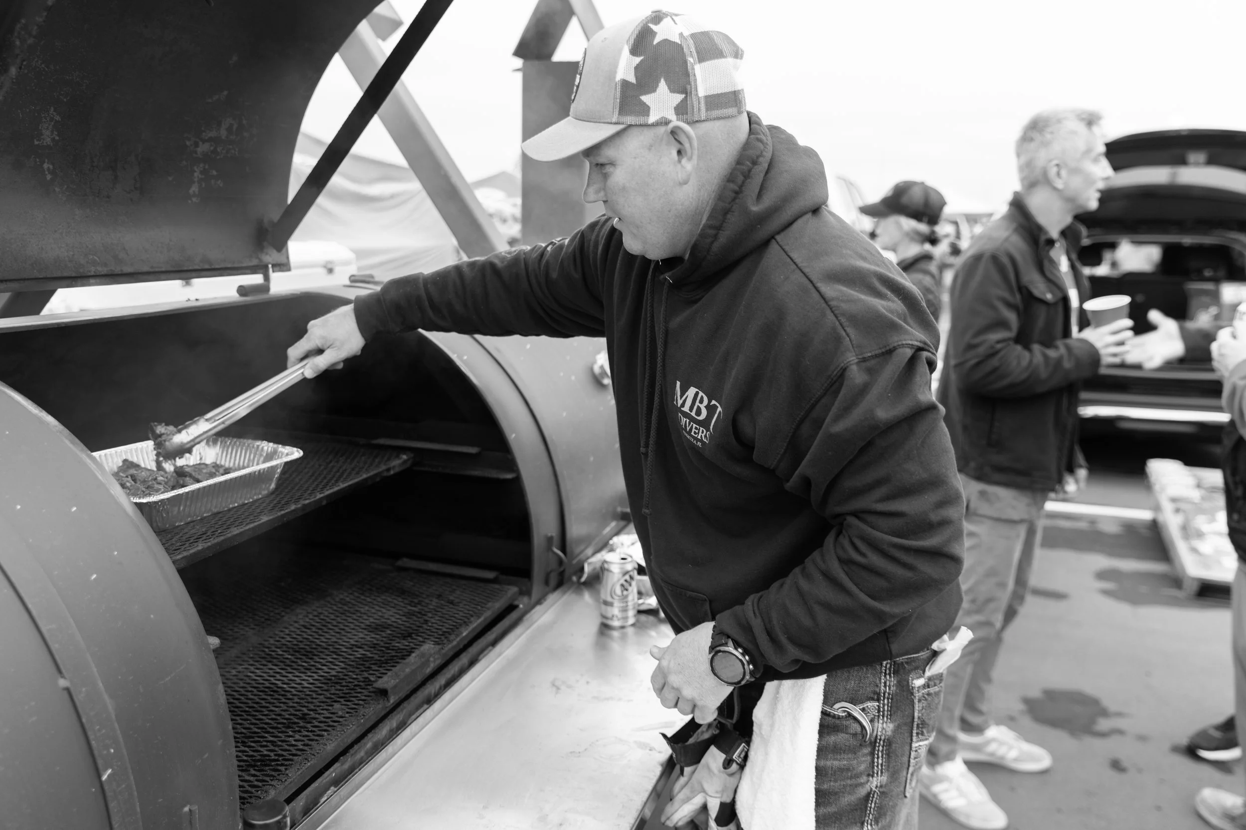 A man wearing a hoodie and a cap with stars and stripes is using tongs to place food on a grill. Others are in the background, some holding cups, around an outdoor event.