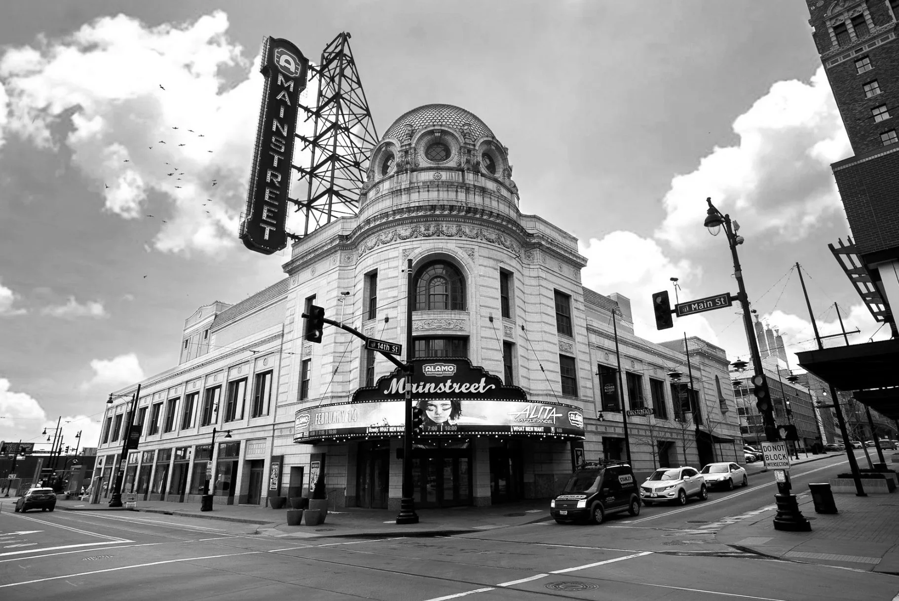 Black and white photo of a historic building at the corner of Main Street and 14th Street, featuring a dome and neon signs for Munst Street and Alamo theater, with parked cars and street signs
