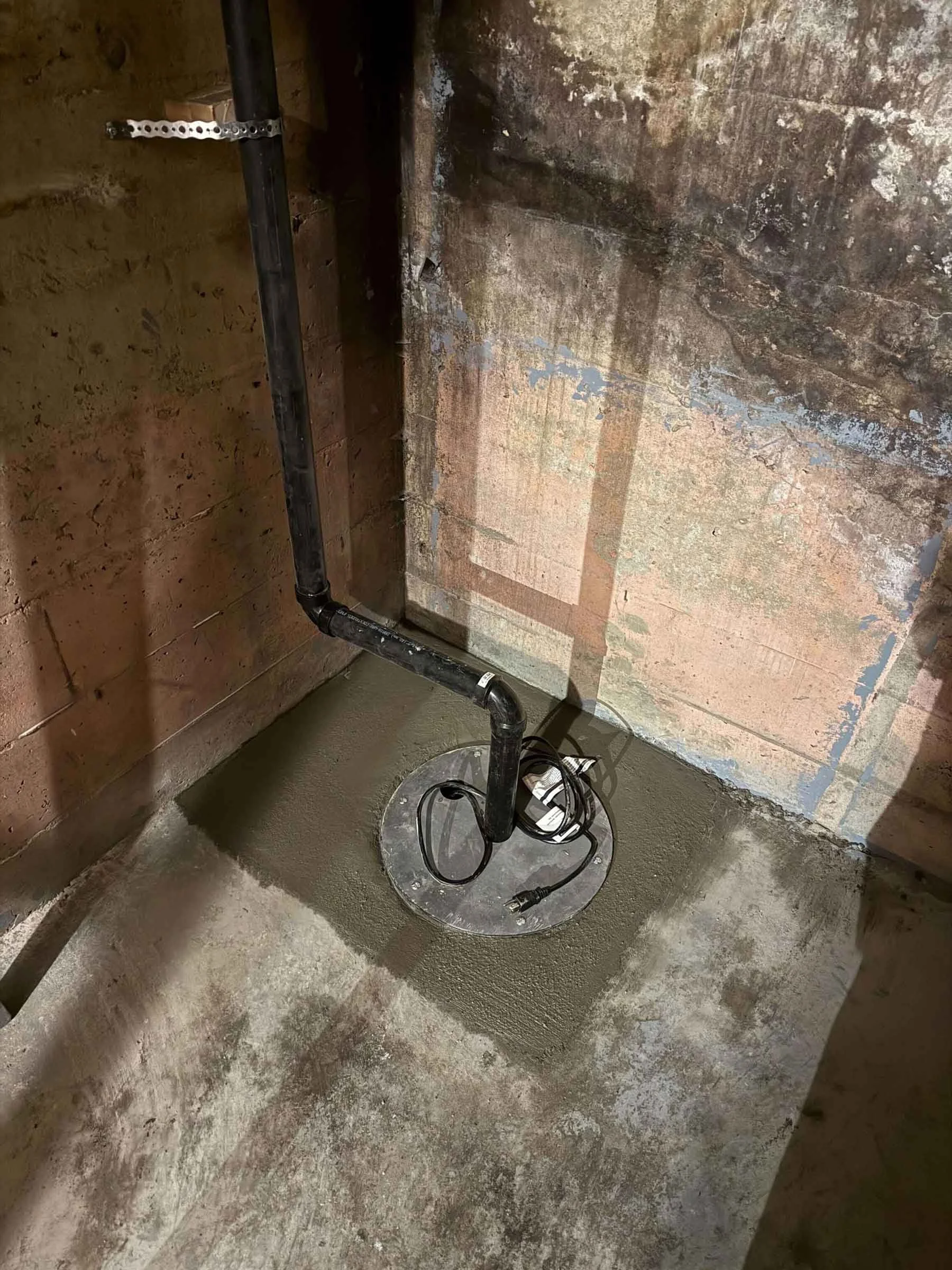 Sump pump and basin installed in basement corner, connected to discharge piping for groundwater management.