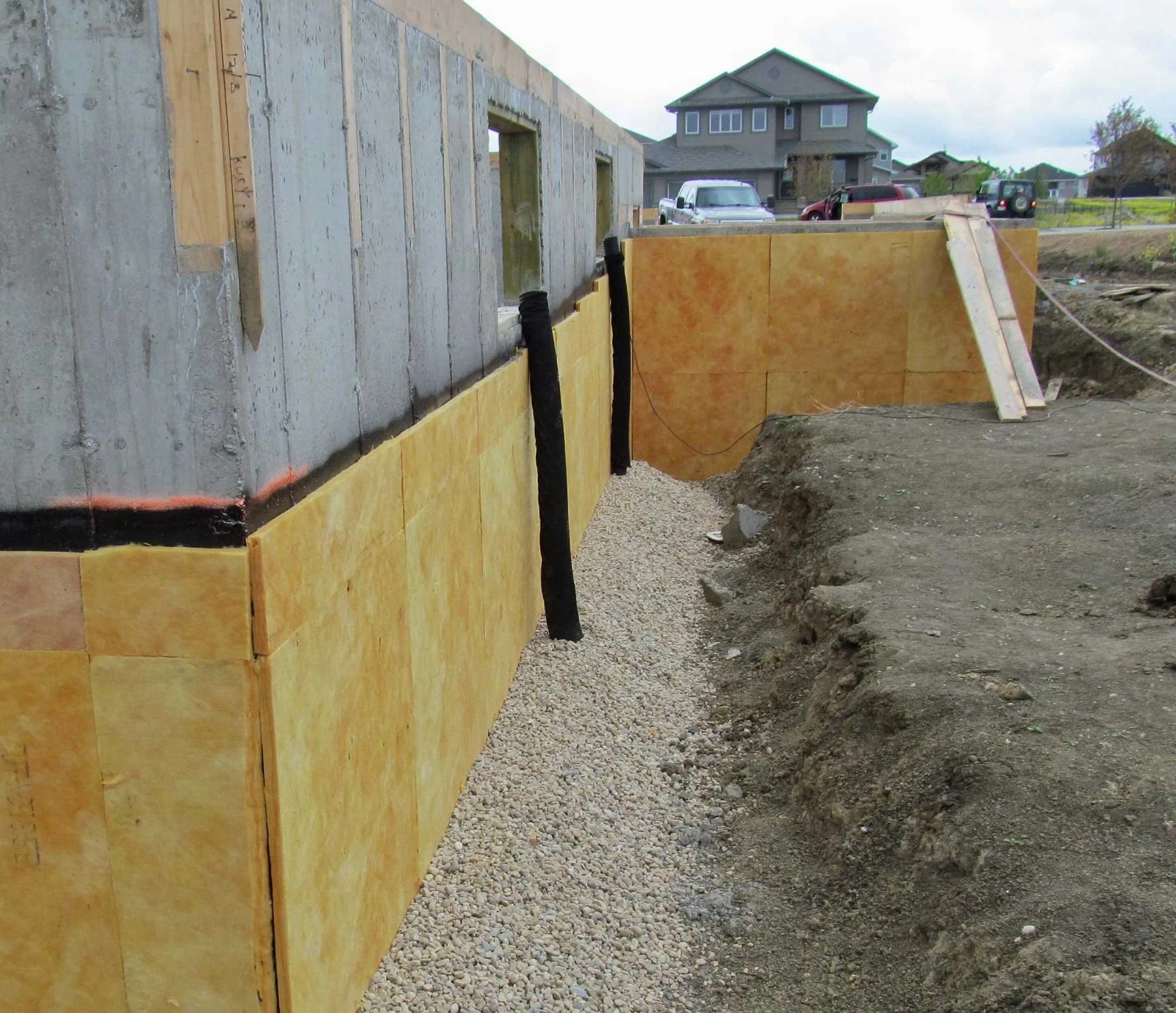 New construction foundation waterproofing with rigid insulation, drainage membrane, and washed rock backfill ready for weeping tile installation.