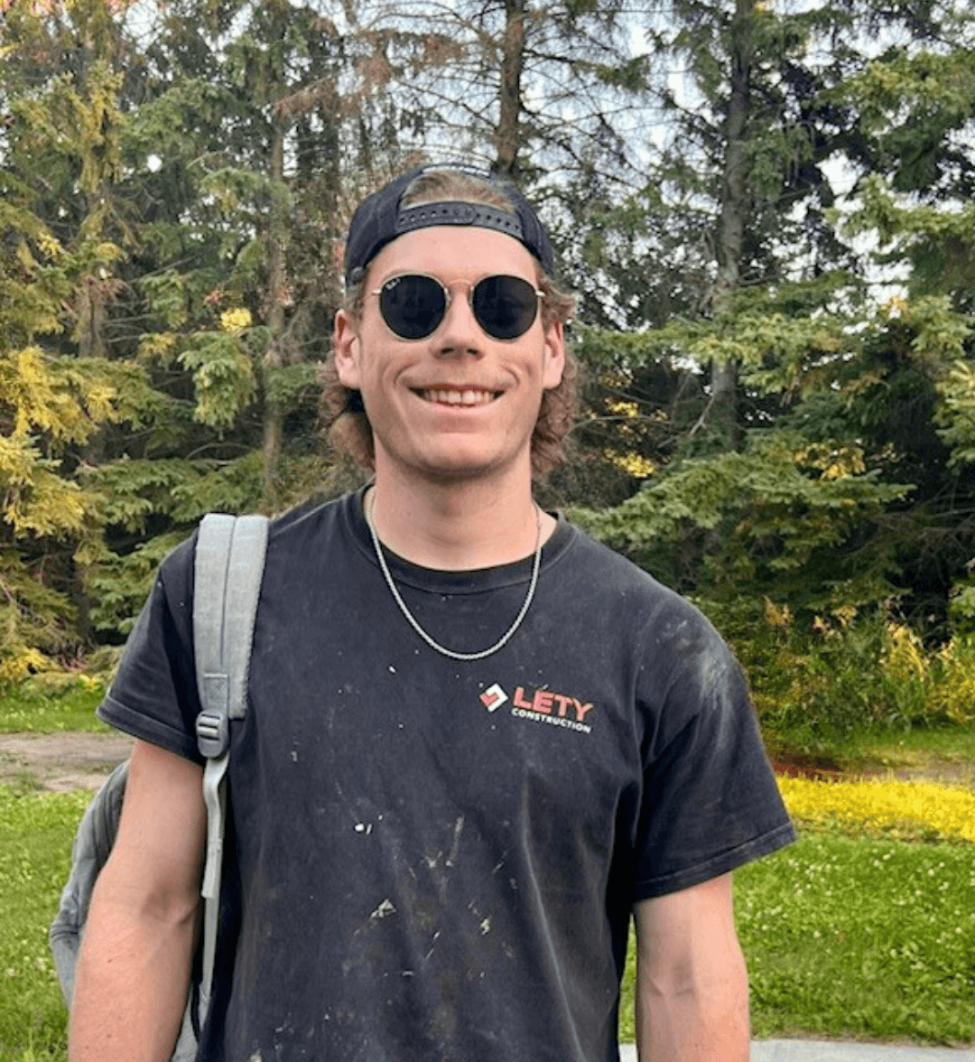 Ty Sutherland, Labourer & Shop Hand, outdoors in front of trees, wearing sunglasses, a backwards cap, a black t-shirt with the logo 'Léty Construction', a silver chain necklace, and carrying a backpack, smiling.