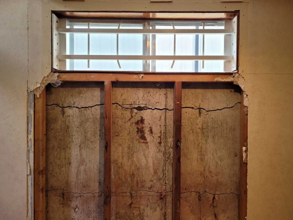 Image shows a wall with a window high up, revealing a building or structure outside. The wall is partially exposed, with drywall removed to show the wooden studs and cracks in the plaster or drywall behind it.