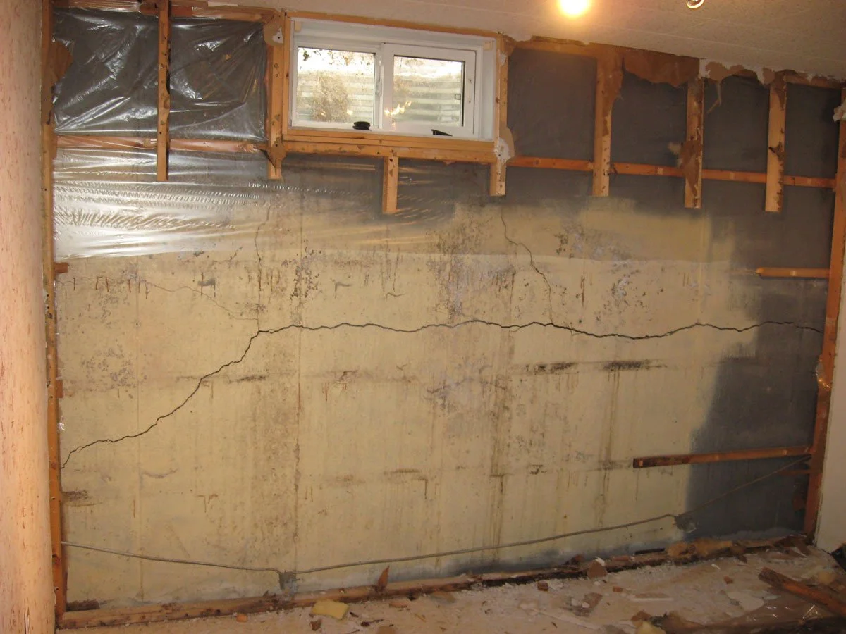 Interior view of a wall with cracks, exposed wooden studs, insulation, and vapor barrier, indicating ongoing renovation or damage repair.