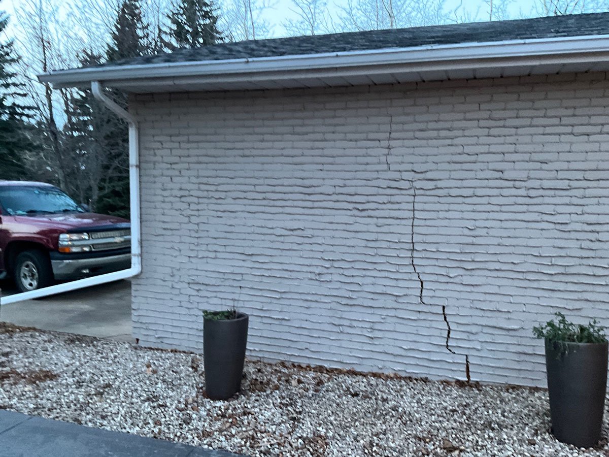 A white brick wall with a large vertical crack running down the middle, two black planters with plants in front, and a vehicle parked beside the wall, with a background of trees and a cloudy sky.