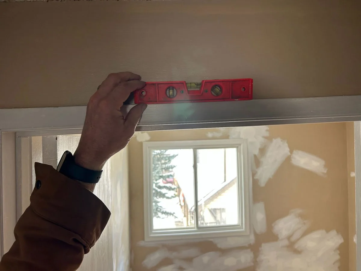 A person using a red level tool to ensure a door frame is straight inside a room under construction or renovation, with a window and drywall visible in the background.