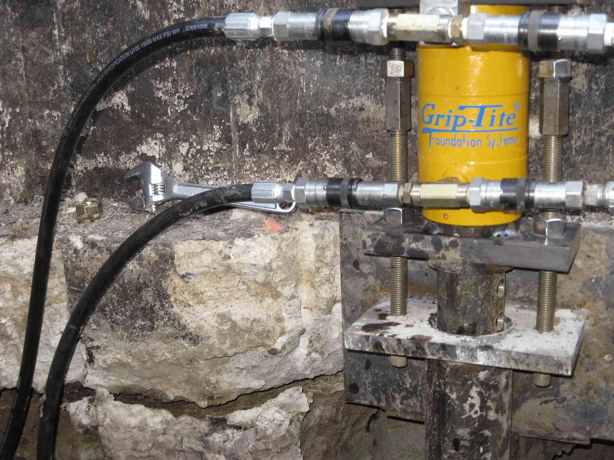Close-up of a mechanical setup with metal pipes, fittings, a yellow Grip-Tite foundation system, and a wrench, mounted on a rough concrete wall.
