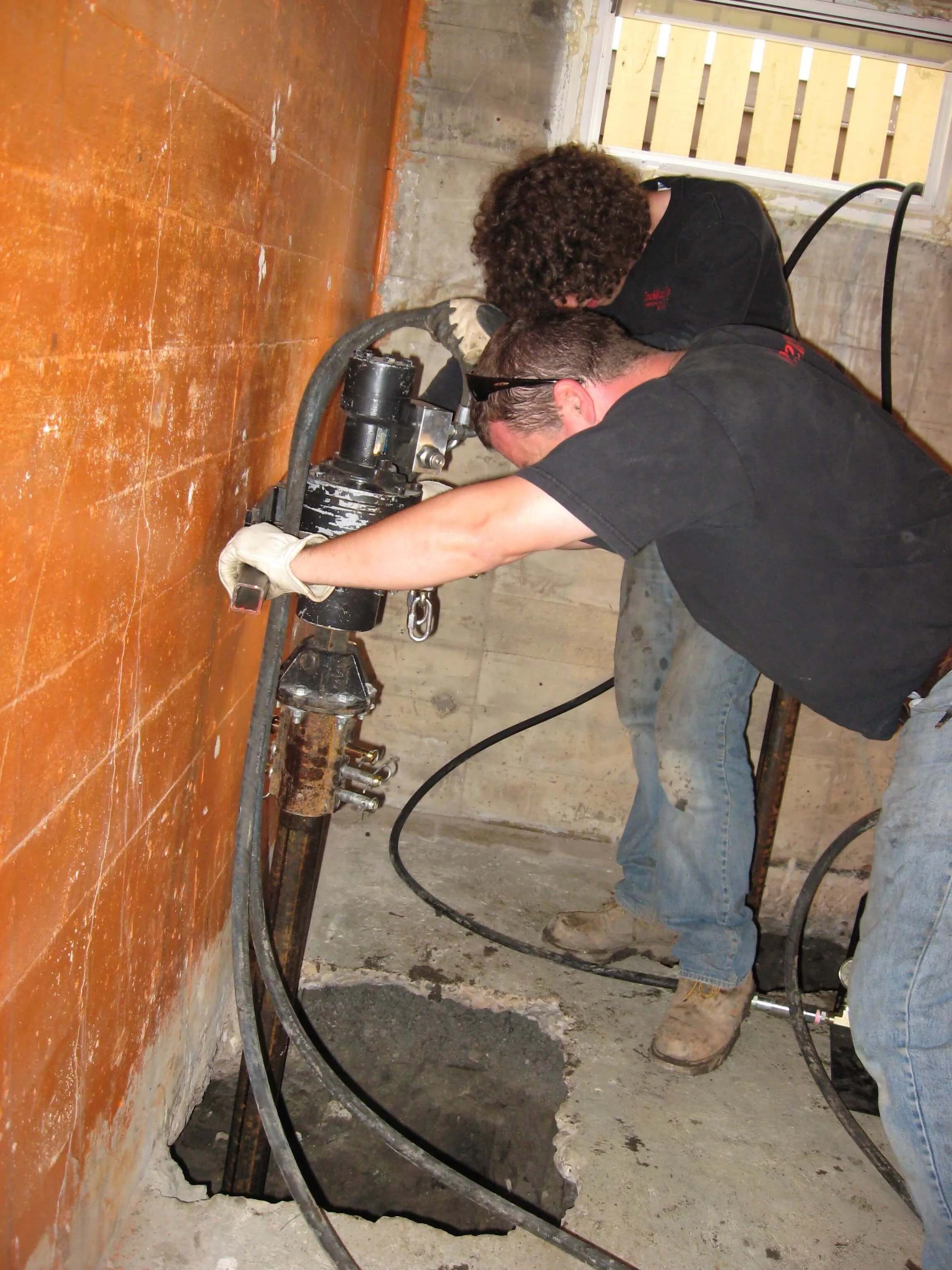 Two workers are installing or repairing a foundation or concrete wall, using specialized equipment and wearing gloves in a construction setting.