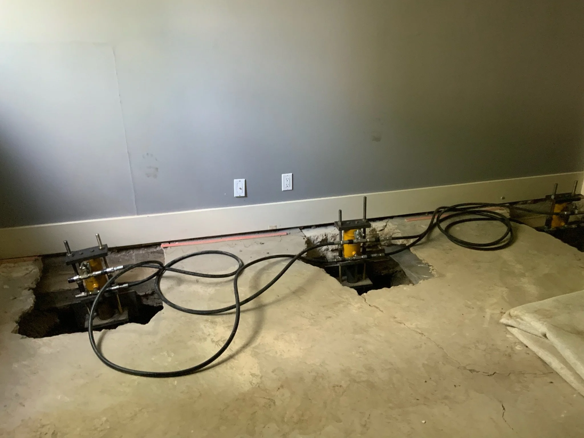 Room with exposed flooring, cut holes in the floor, and machinery with hydraulic hoses, likely for plumbing or construction work.