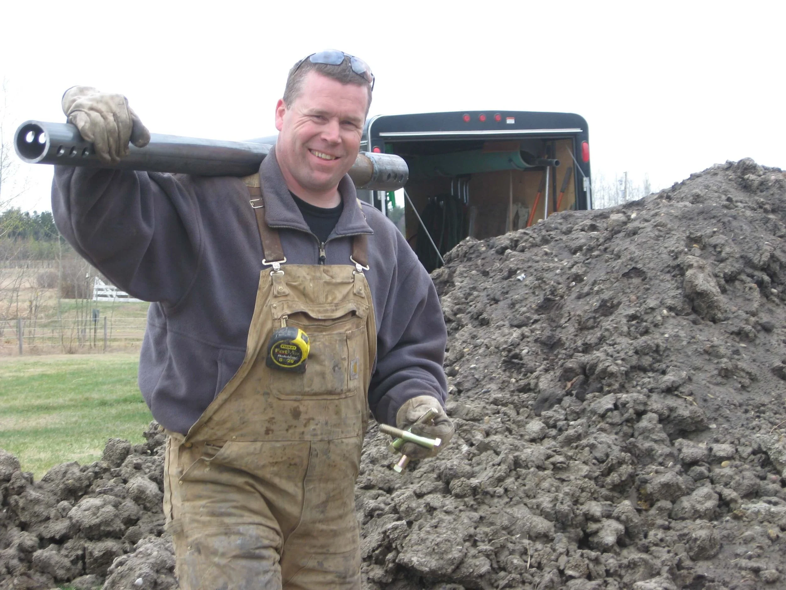 Wes Sutherland smiling outdoors, holding a shovel over his shoulder, standing in front of a large pile of dirt, with a trailer in the background.