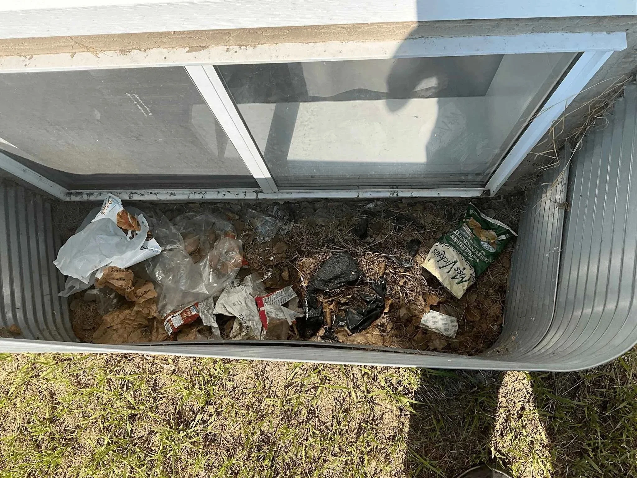 Window well filled with leaves and debris, illustrating the need for regular cleaning to prevent drainage issues.