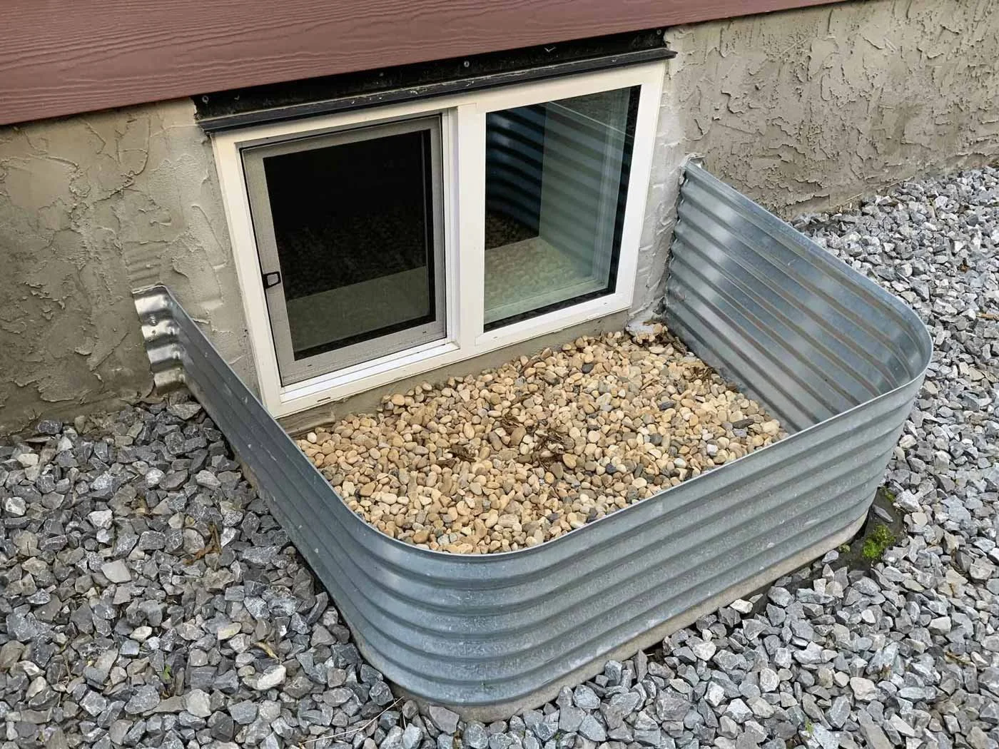 A metal window well with gravel inside, surrounding a basement window on an exterior wall of a house, with stone gravel spread on the ground outside.