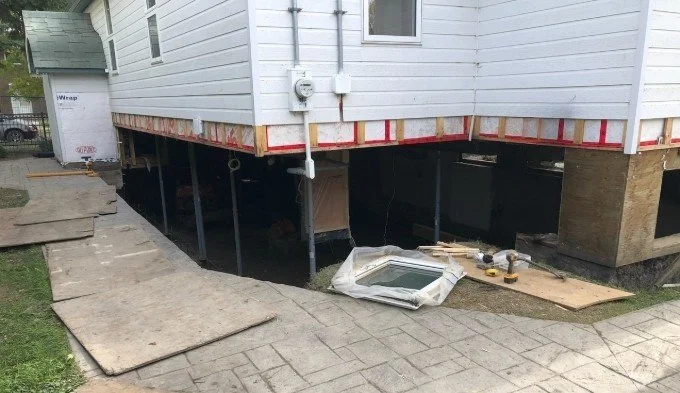 Home supported on temporary bracing during foundation replacement, with new waterproofing membrane applied to the exterior walls.