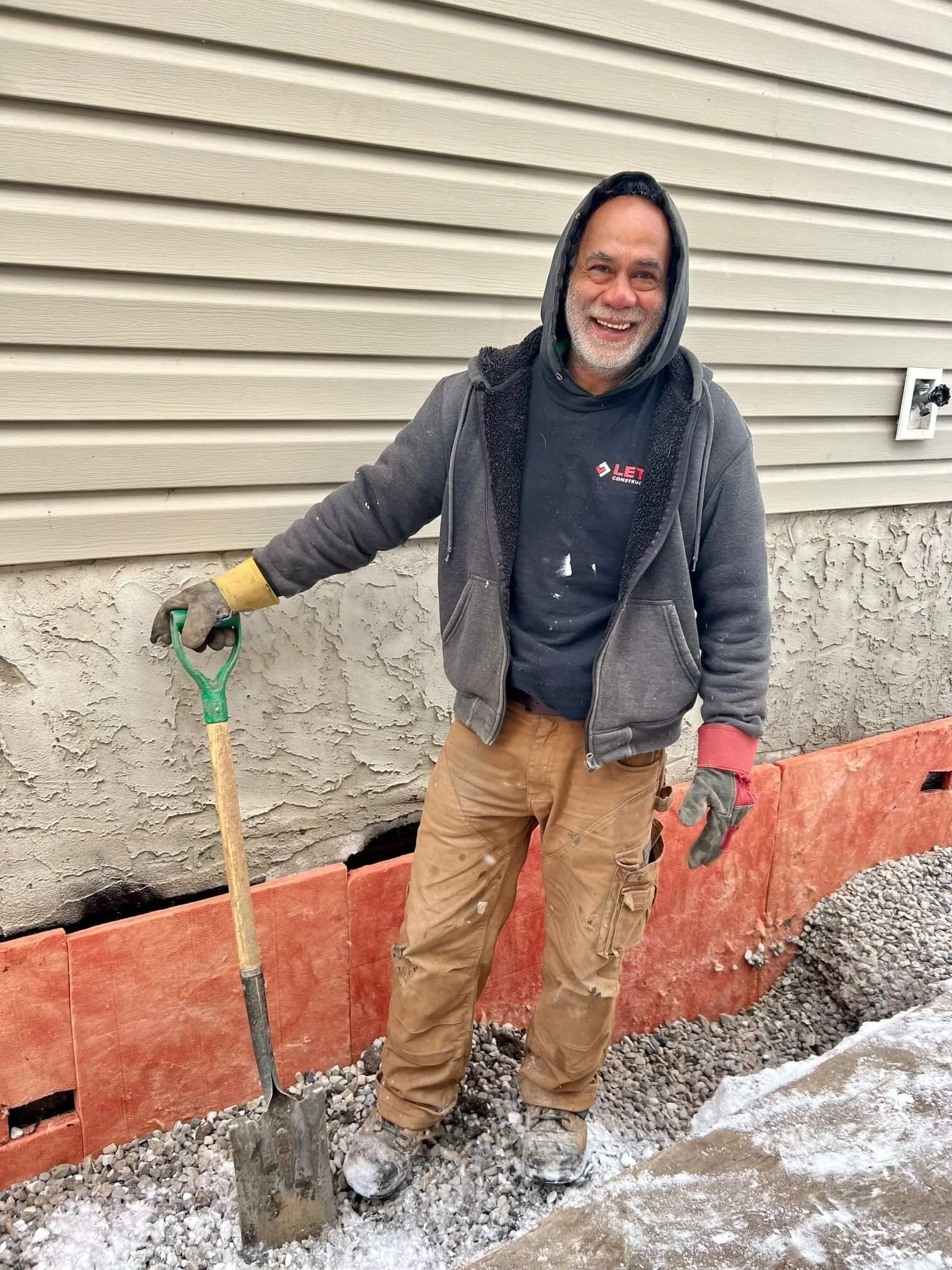 Carl Maripain, Senior Labourer, smiling outdoors on a construction site, holding a shovel, wearing work gloves, a hoodie, and work clothes, with a building wall and some gravel in the background.