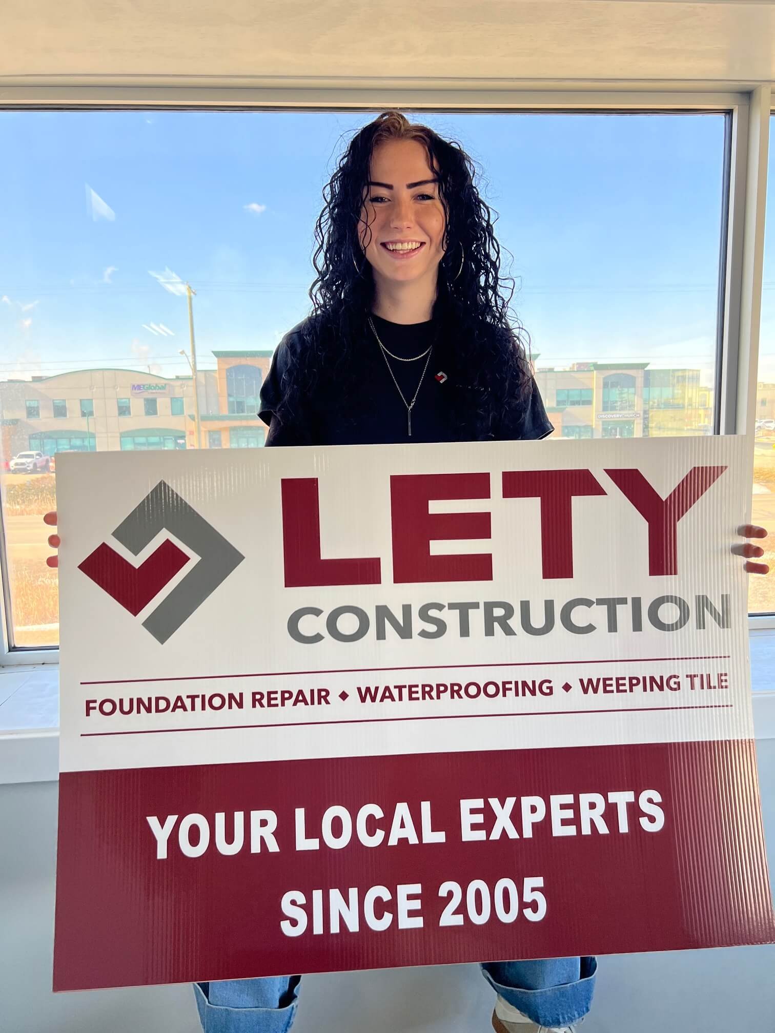 Leah Sutherland inside in front of a large window, holding a sign that reads 'Lety Construction,' with services listed including foundation repair, waterproofing, and weeping tile. 