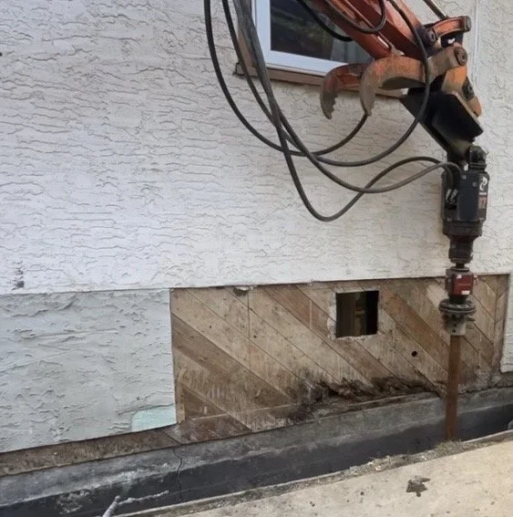 Crew members driving a helical pier into the ground using excavator-mounted equipment during a residential foundation underpinning project.