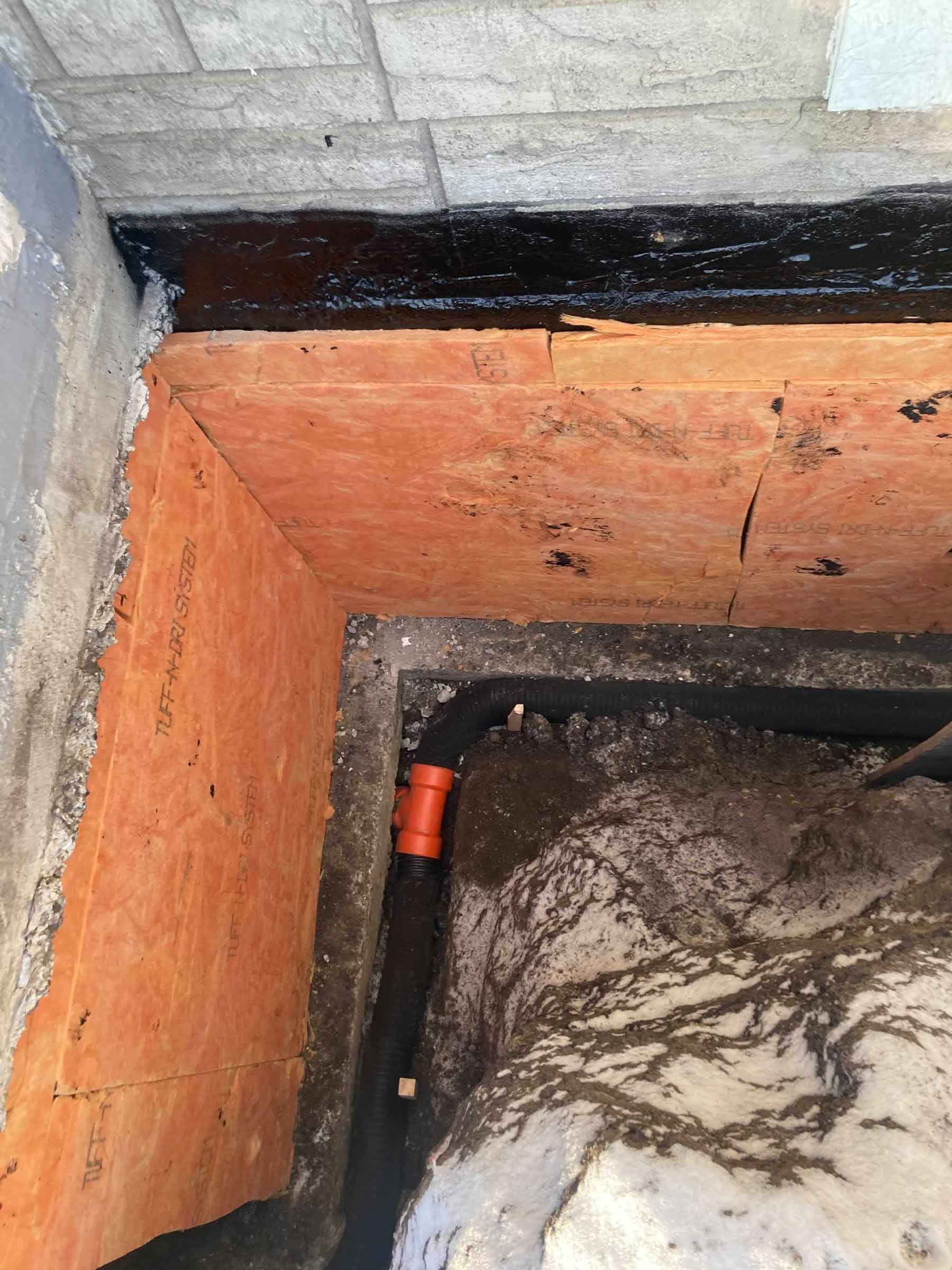 Exterior foundation excavation showing waterproofing membrane, rigid insulation, and sump pump discharge connection.