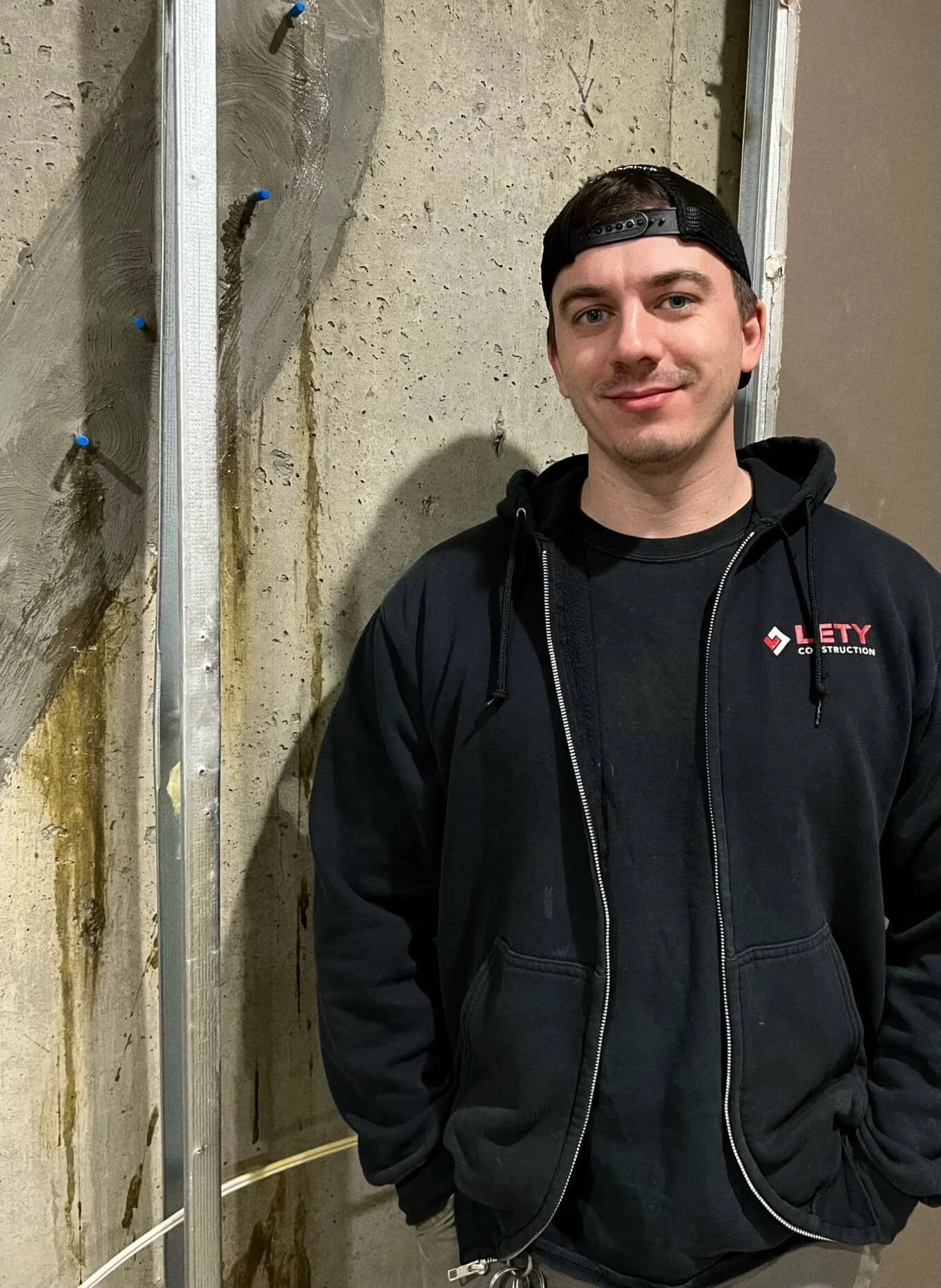 Vadym Robaschuk, Senior Labourer, wearing a black hoodie and cap, standing indoors against a concrete wall, smiling at the camera.