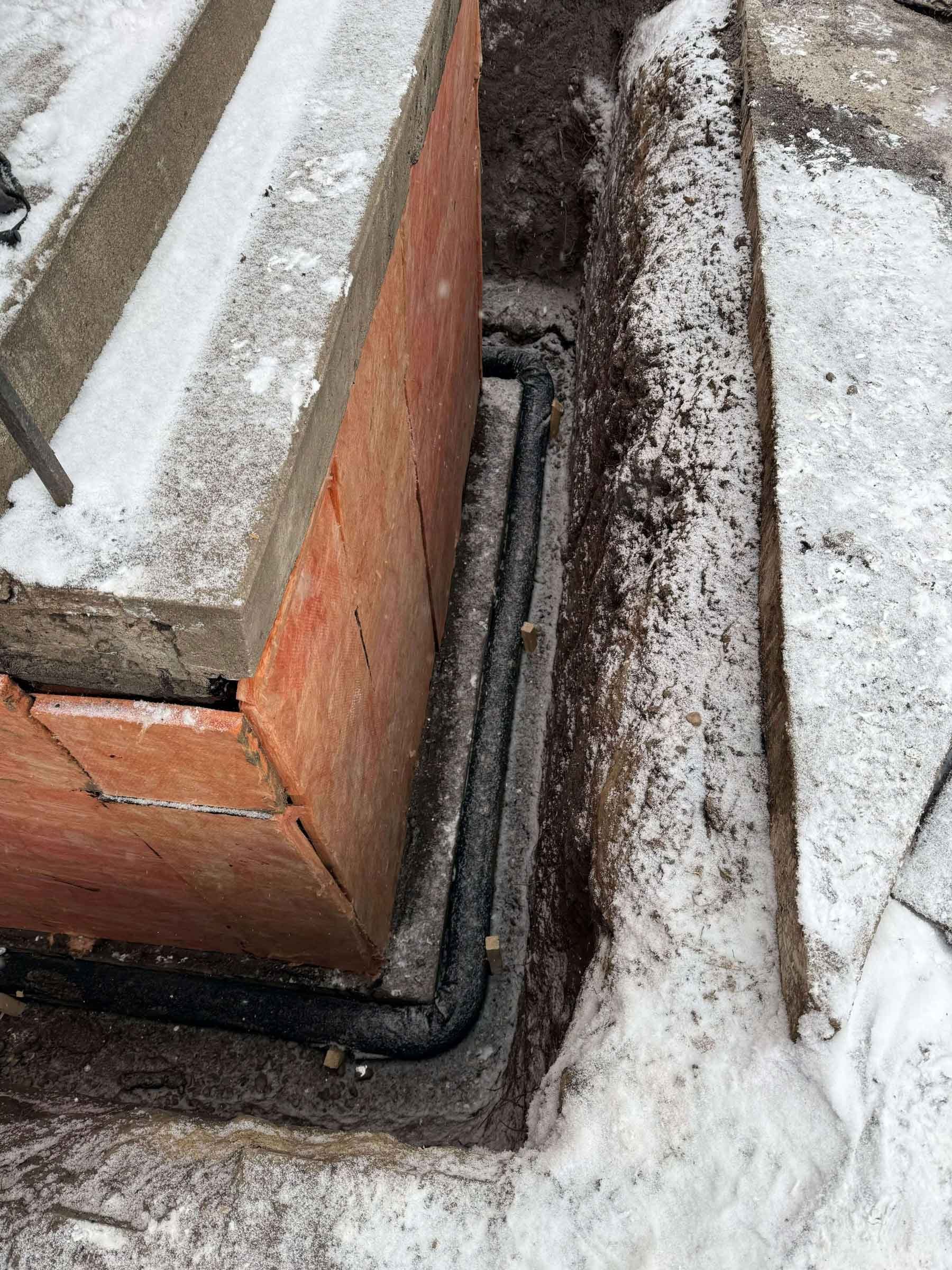 Exterior weeping tile installation at a foundation corner, with new drainage pipe placed in the excavated trench and rigid insulation protecting the foundation wall.