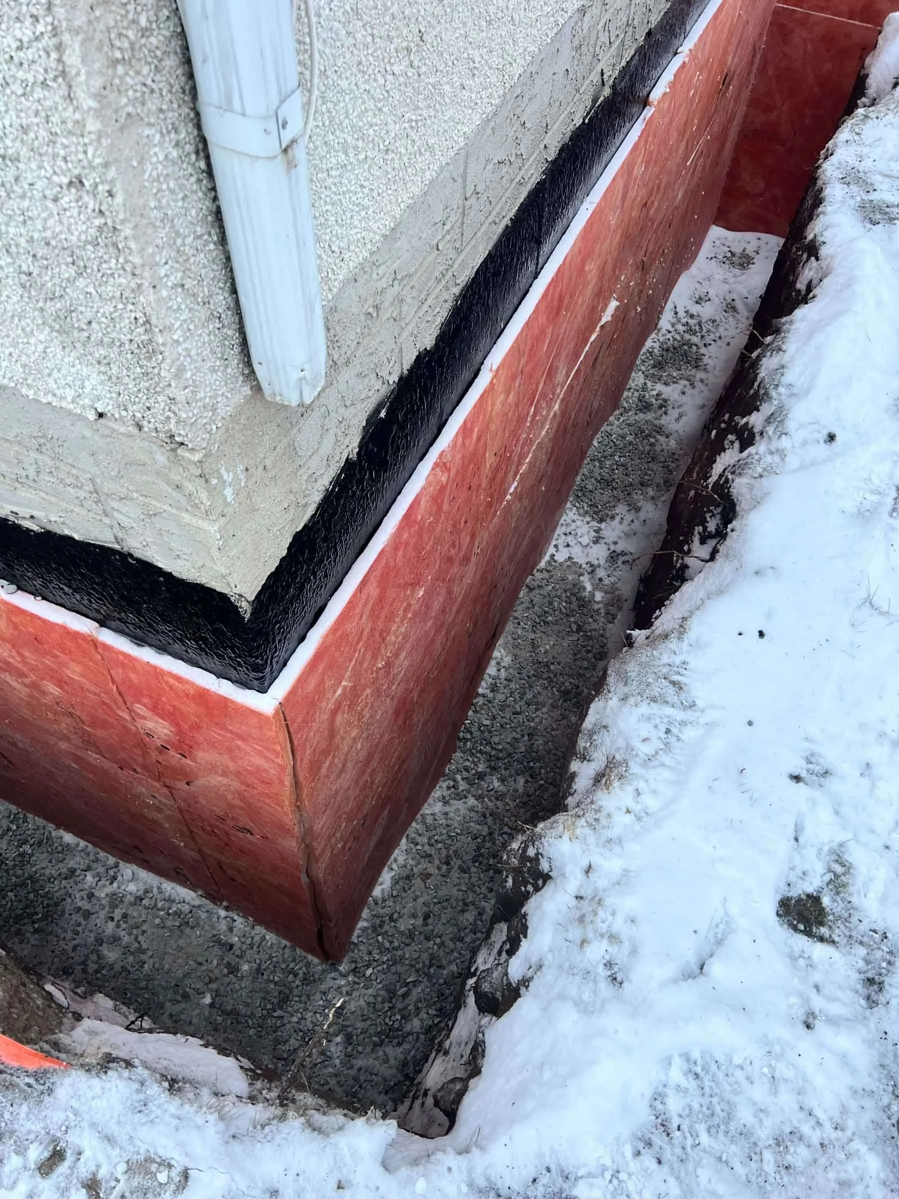 Exterior foundation corner with waterproofing membrane, insulation board, and downspout drainage integration.