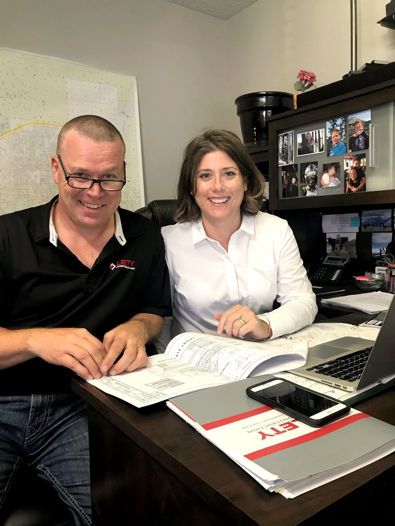 Wes and Rachelle Sutherland, Owners of Lety Construction, sitting at a desk with documents, a laptop, and a smartphone, in an office with photo collage on the wall.
