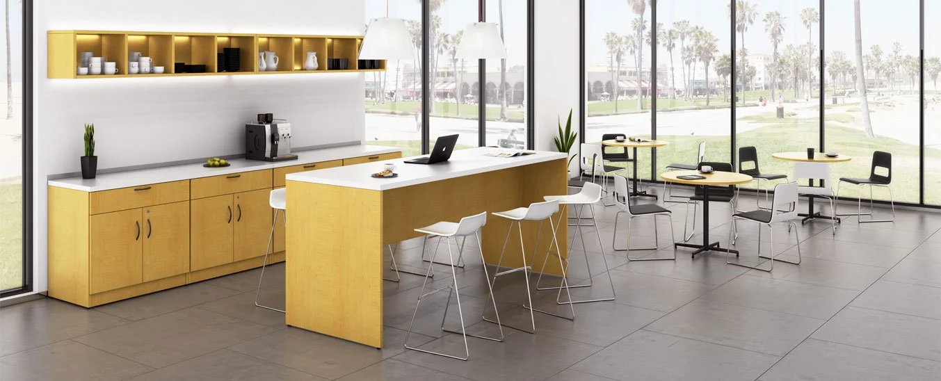 Modern office break room with yellow cabinets, a kitchen island, and seating area with round tables, white and black chairs, large windows, and a view of a park.