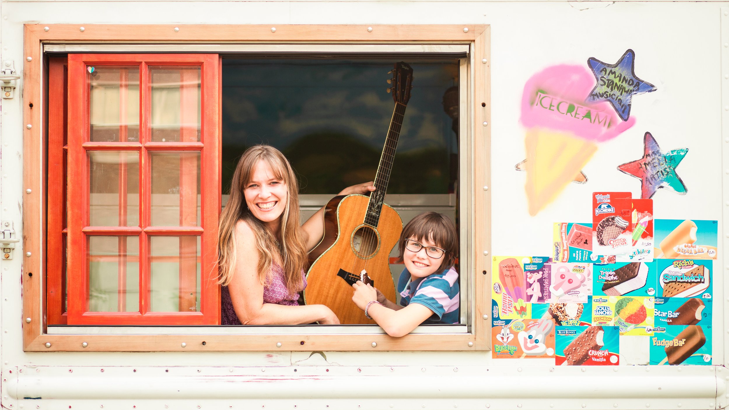 A smiling woman with long blonde hair and a young boy with glasses and dark hair are inside a small ice cream stand, with a red window frame. The woman is holding a guitar, and the boy is holding a piece of ice cream cake. The stand is decorated with colorful ice cream-themed paintings and advertisements.