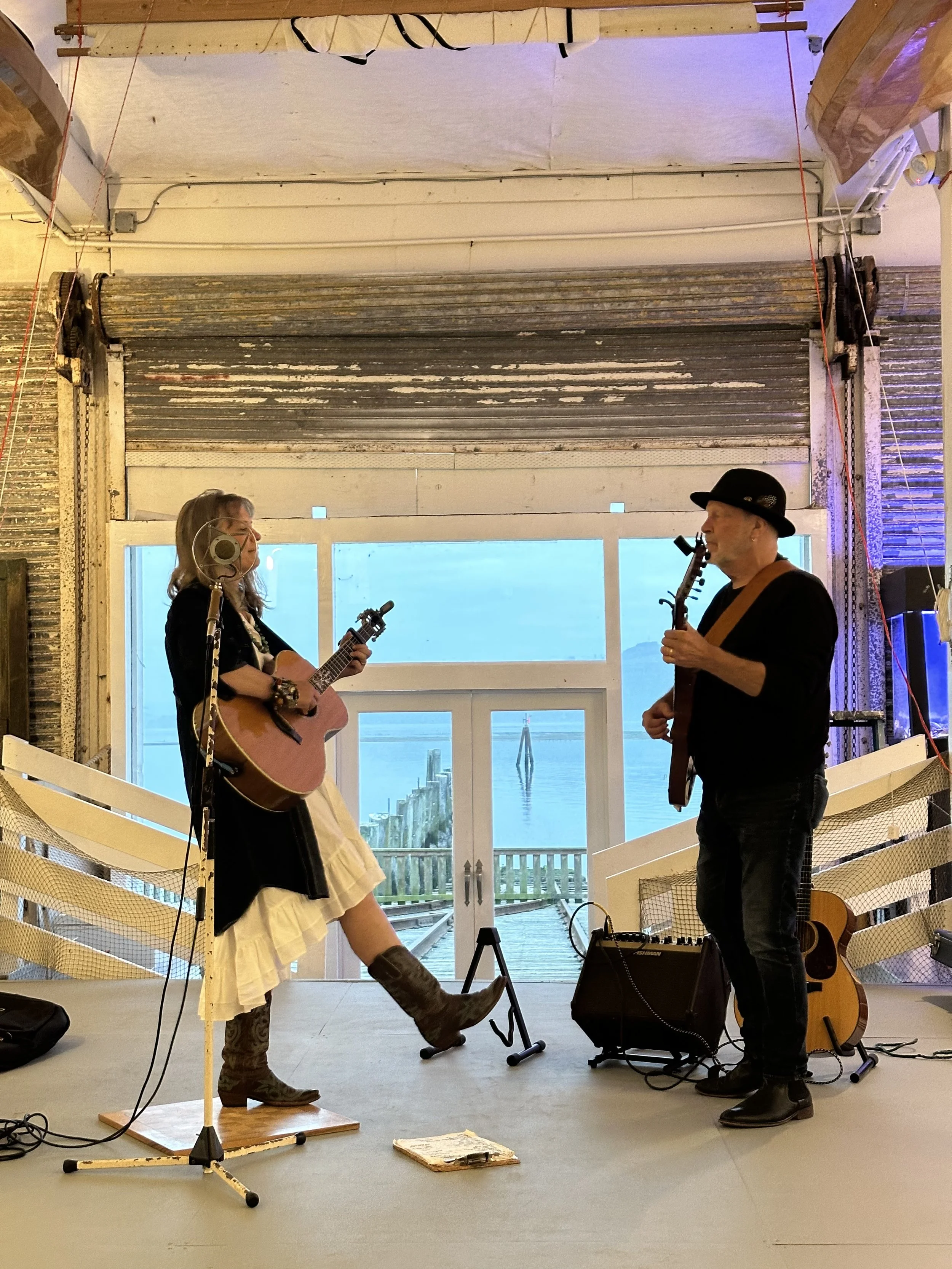 Two musicians perform indoors near large windows overlooking a body of water. One is a woman with long hair, playing a guitar and wearing a black jacket, white skirt, and knee-high boots. The other is a man with a beard, wearing a hat, holding a guitar, and smoking a pipe. There are microphone stands, a guitar on a stand, and electronic equipment, with a view of water and mountains through the windows.