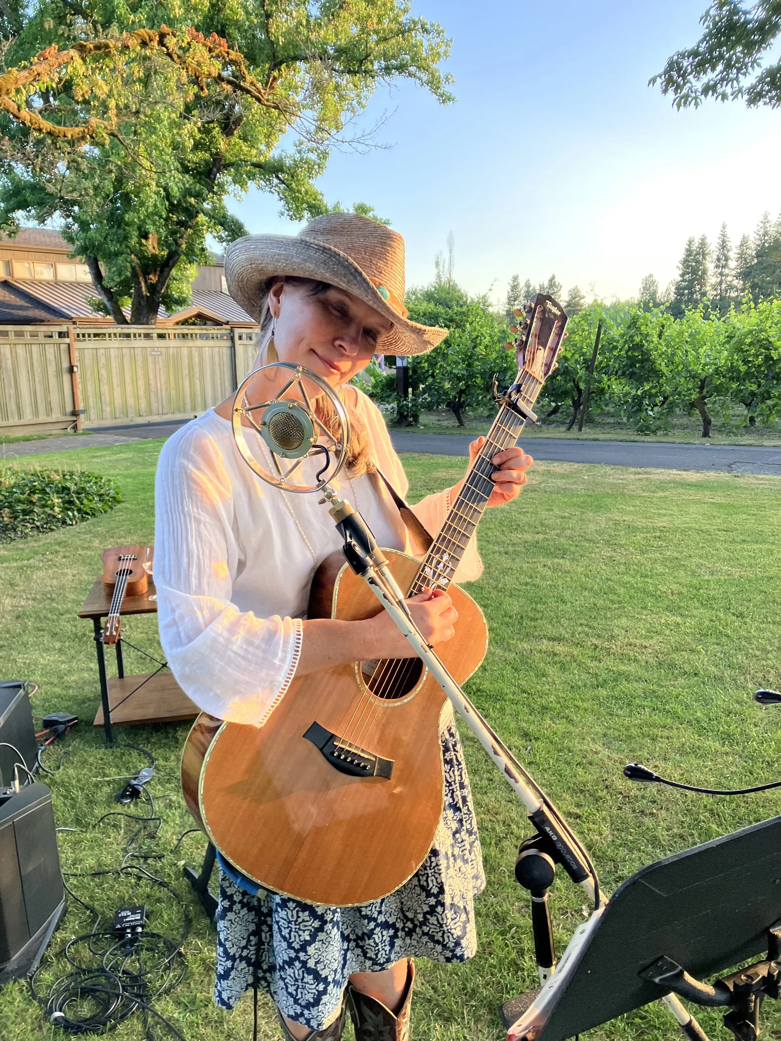 A woman standing outdoors in a green yard, wearing a straw hat, playing an acoustic guitar and singing into a microphone. There is audio equipment and a second guitar on a stand nearby, with a backdrop of trees, a wooden fence, and a house at sunset.