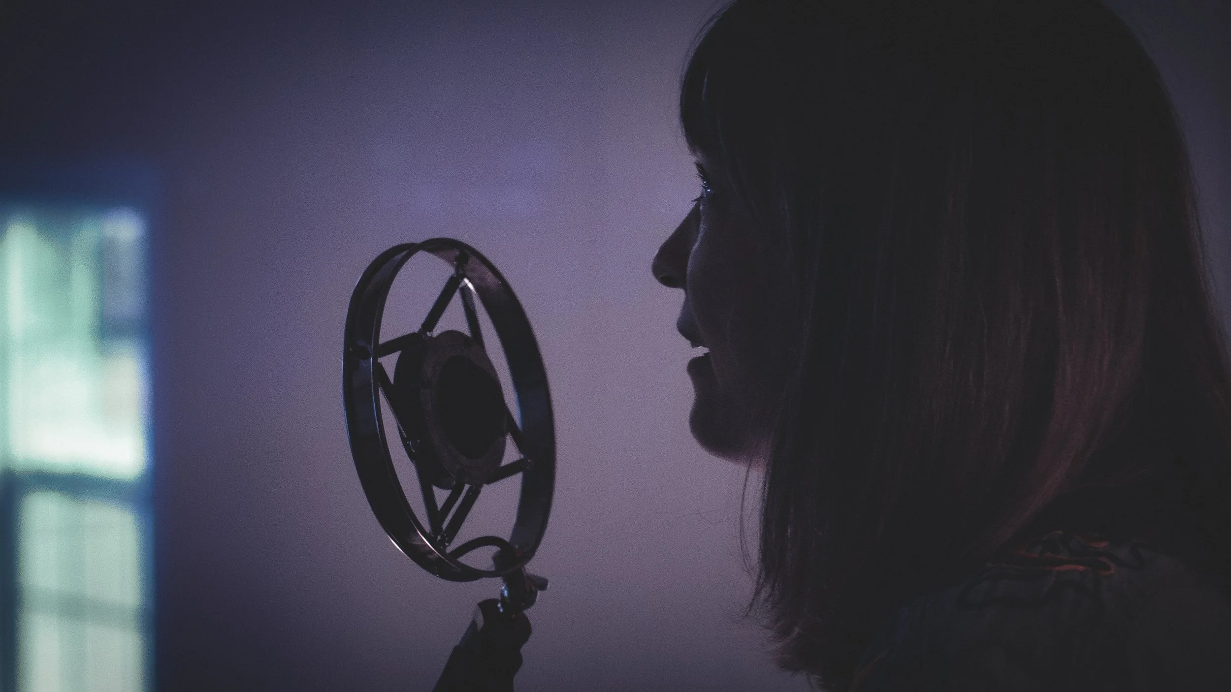 A woman with long hair singing into a microphone in a dark room with a faint glow on her face.