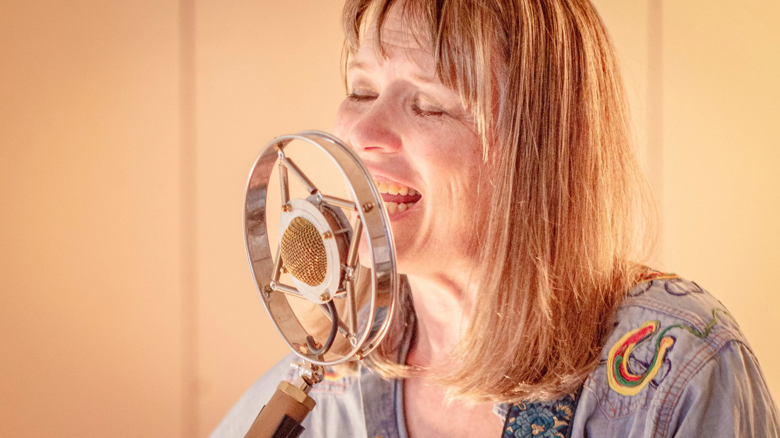 A woman singing into a professional studio microphone with her eyes closed, in a recording studio.