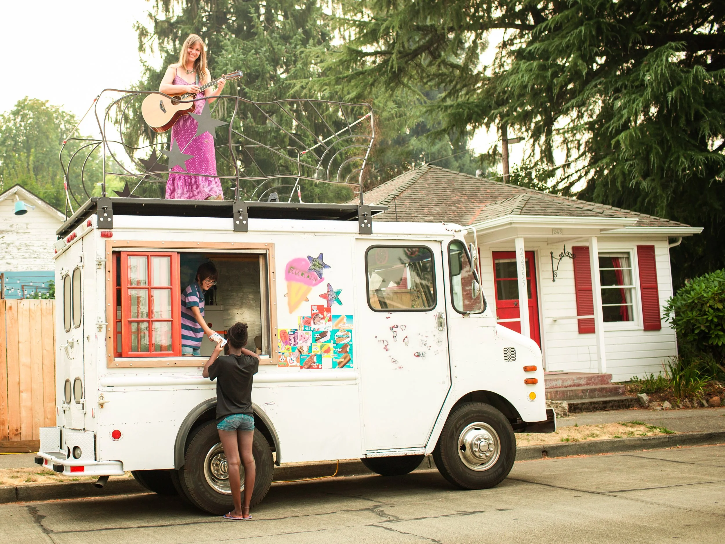 A woman playing guitar on top of a food truck with three stars and ice cream decoration on the side, parked on a residential street with trees and houses in the background.