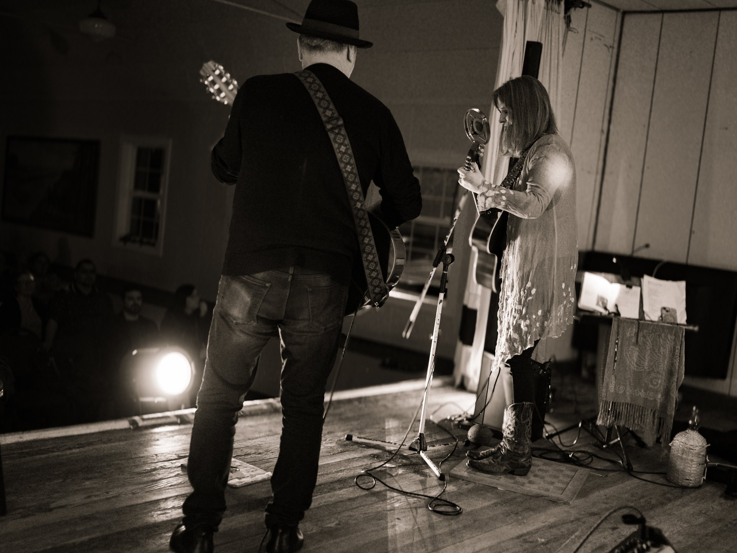 Two musicians performing on a stage, one playing guitar and the other singing into a vintage microphone, with an audience watching attentively.
